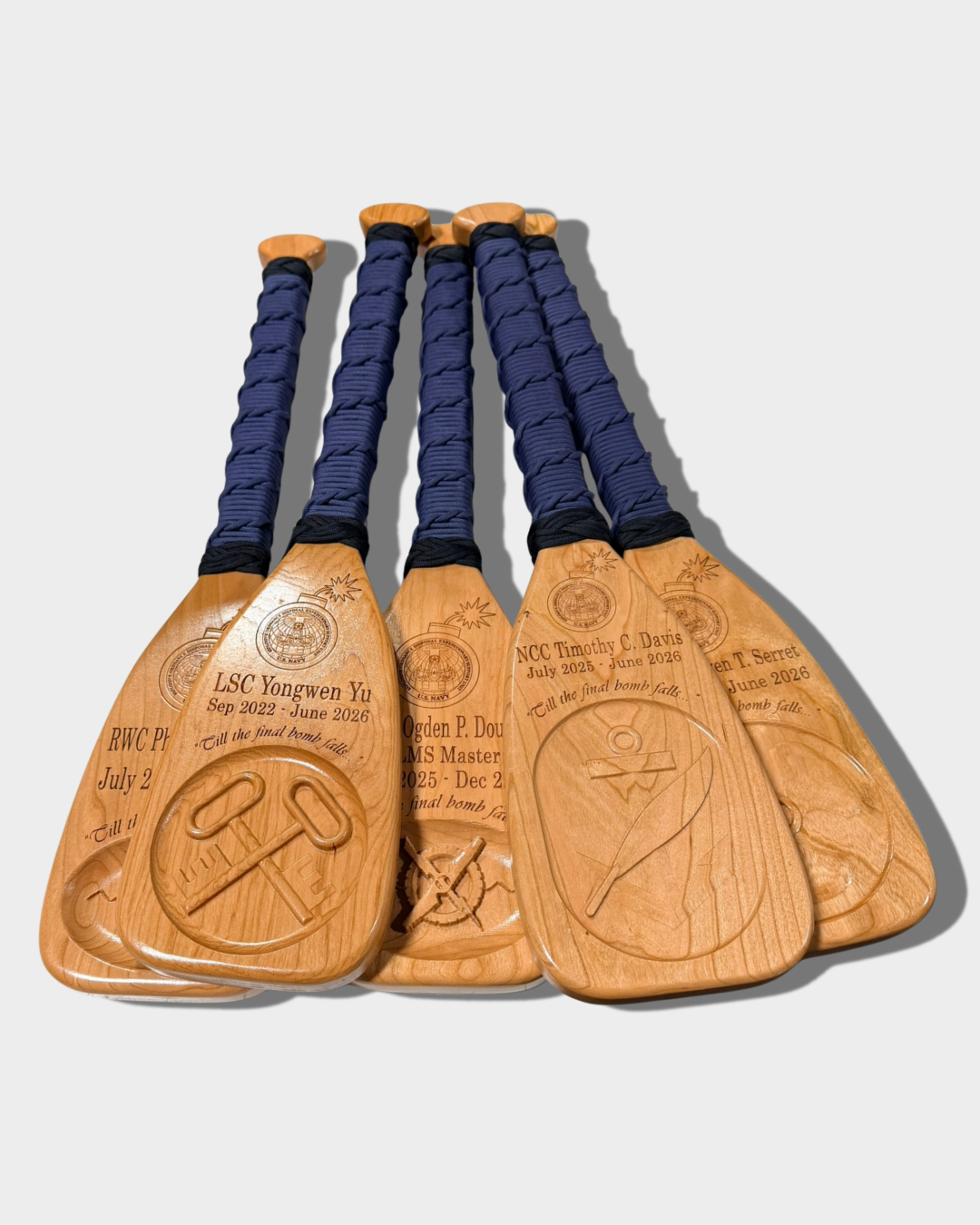 Group of wooden paddles with engraved text and military insignia and symbols, tied with navy blue paracord, lying on a light gray background.