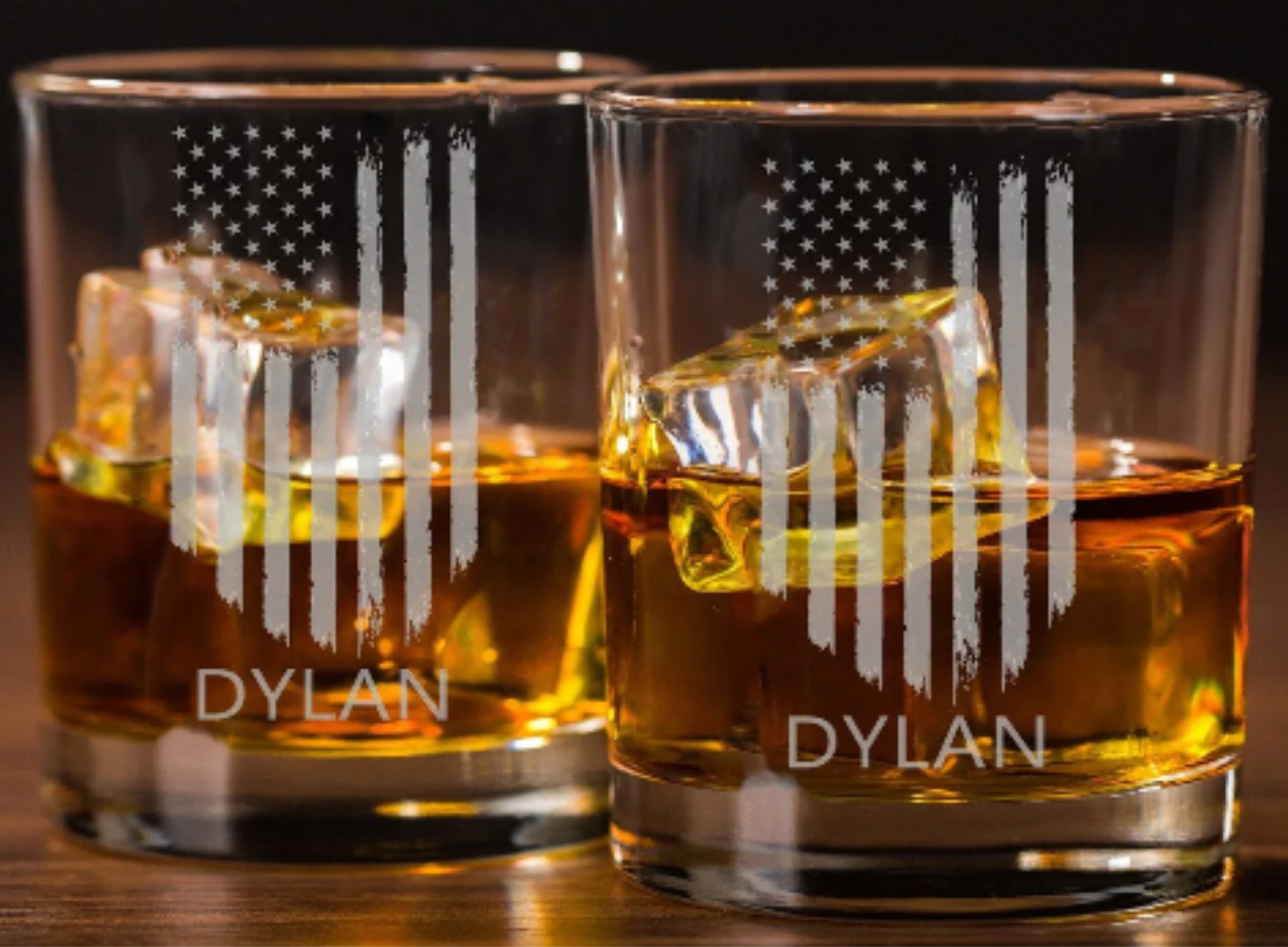 Two glass tumblers with whiskey and ice cubes, glasses have a heart design with an American flag pattern and the name 'Dylan' at the bottom.