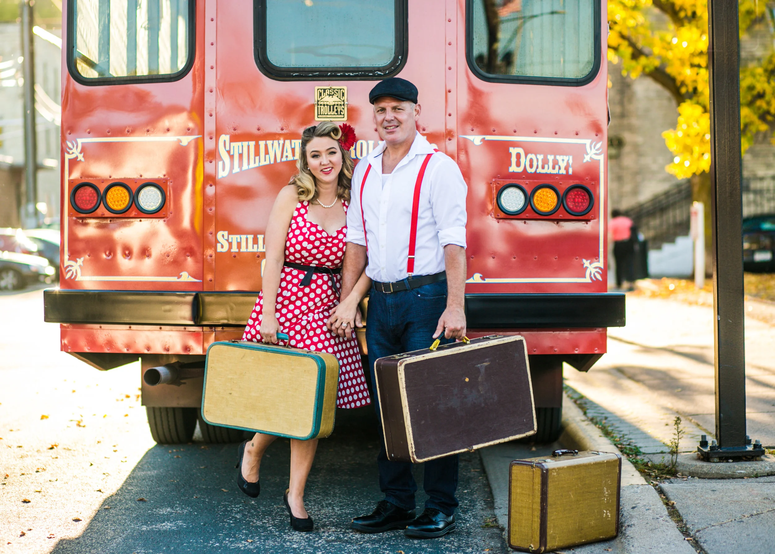 A couple poses with suitcases in front of the Stillwater Trolley during their engagement session with Andrea Wagner Photography