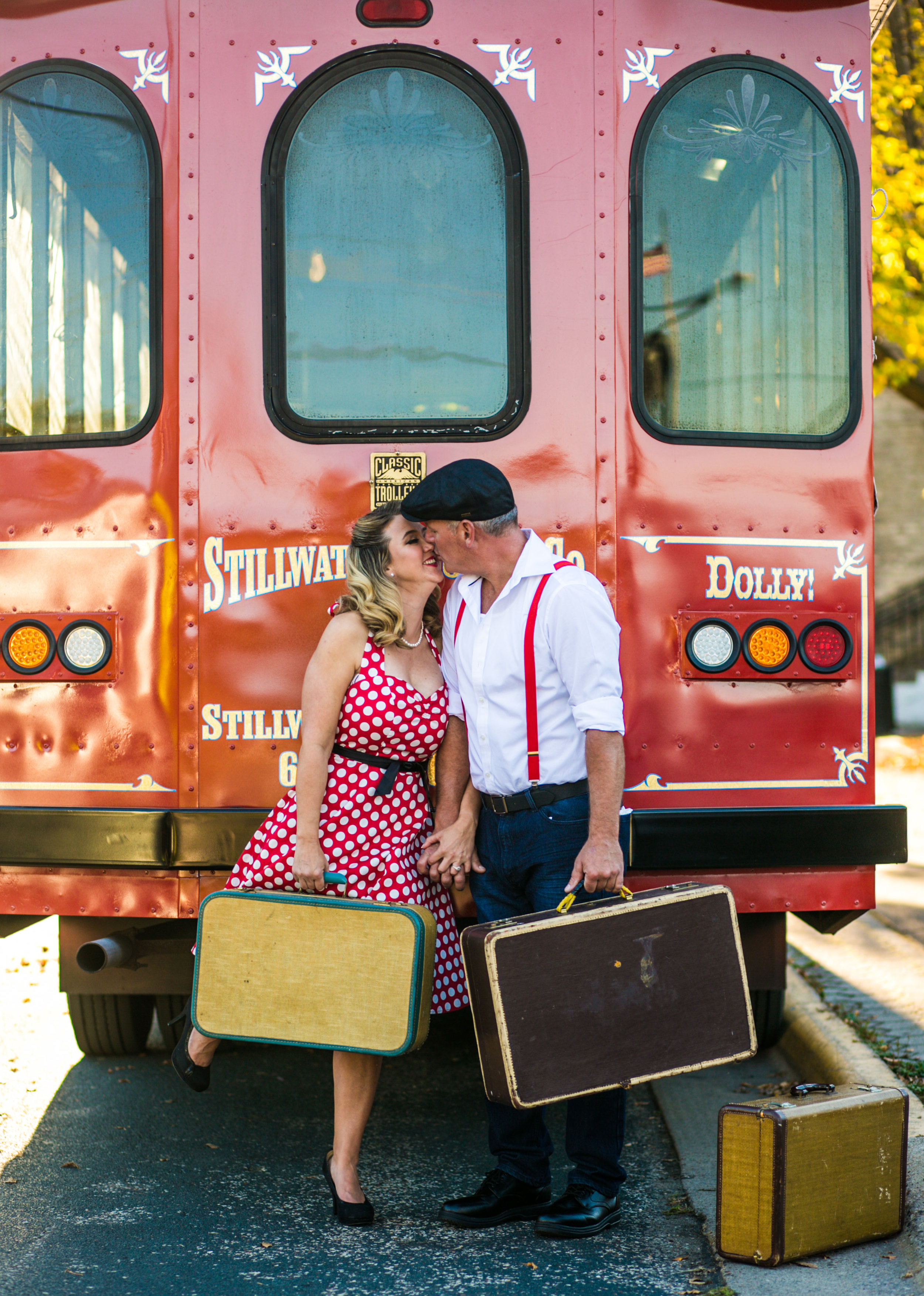  A couple kisses holding suitcases in front of the Stillwater Trolley during their engagement session 