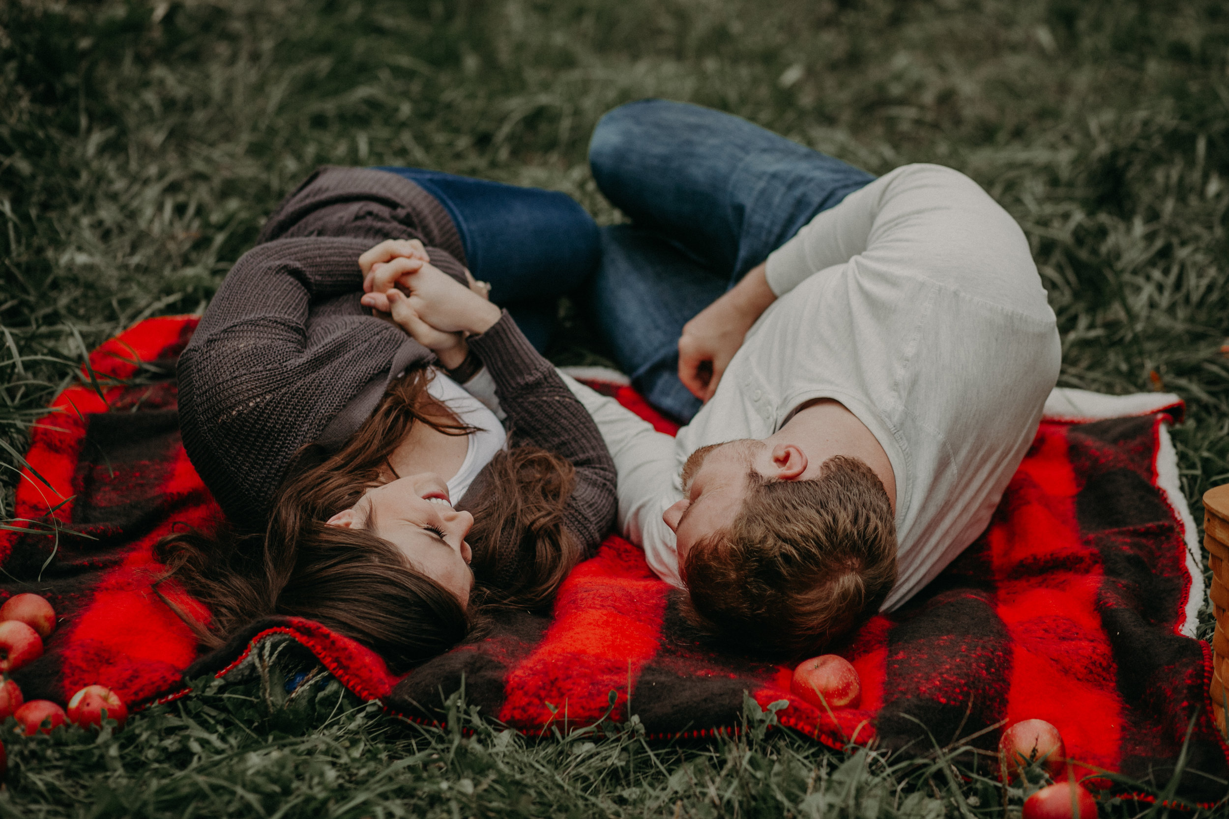  A couple snuggles and holds hands on a plaid red blanket during their engagement session at an apple orchard in the fall 