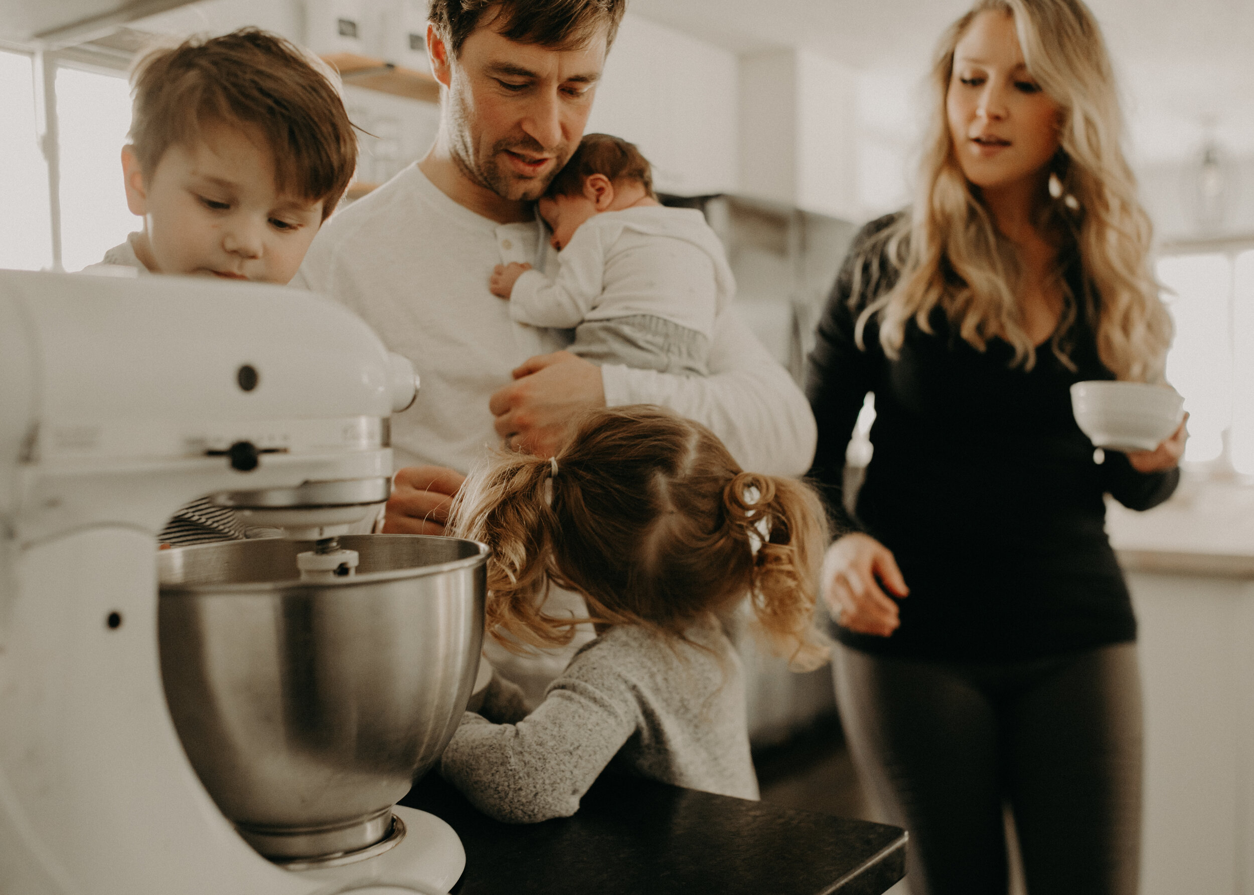  River Falls Family. River Falls Wisconsin. Mother cooking in the kitchen. Lifestyle Newborn Photo session. Wisconsin Newborn Photographer. Wisconsin Lifestyle Newborn Photographer. Lifestyle Newborn In Home Session. Beautiful mom in white making cho