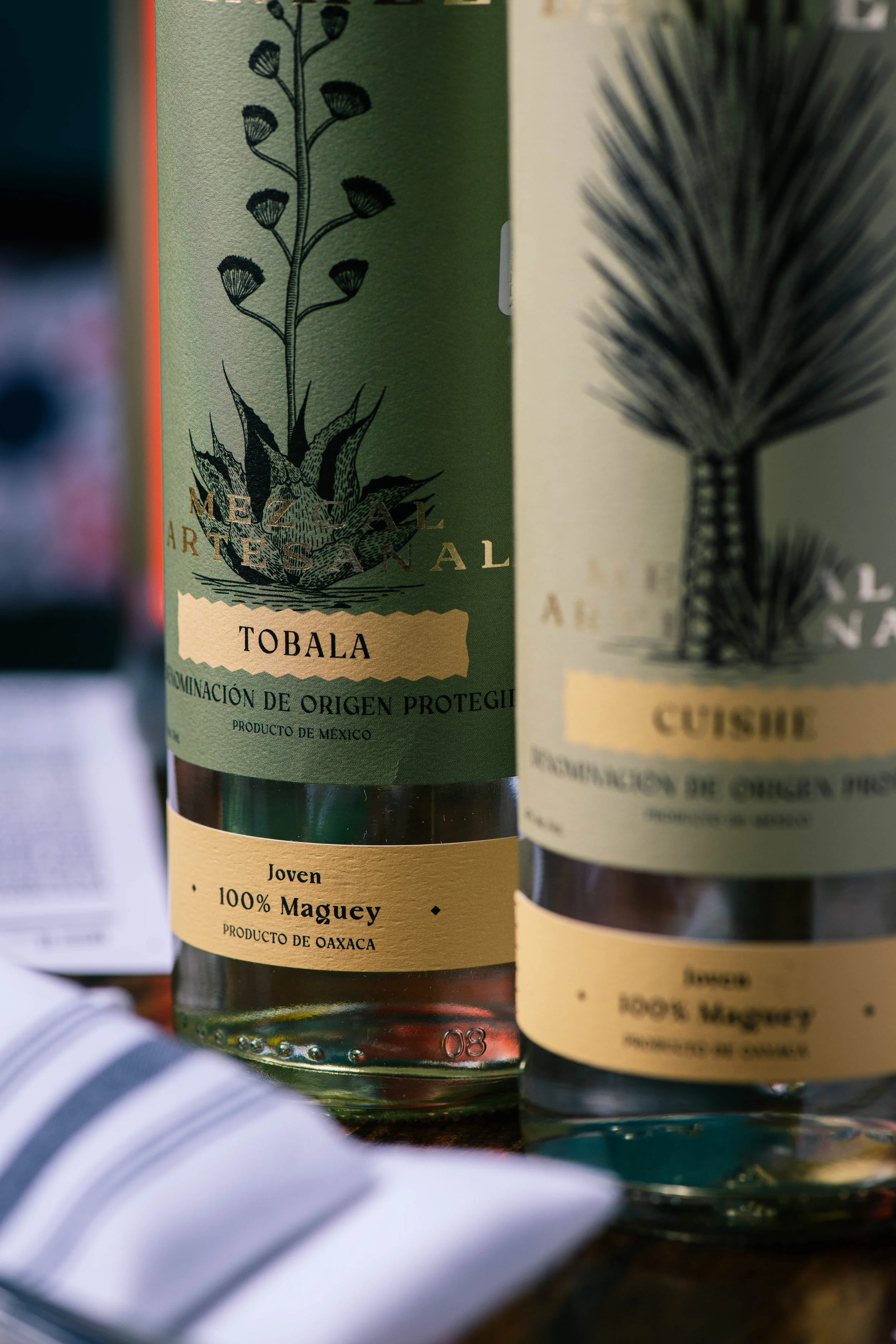 Close-up of two bottles of Mexican agave liquor, showing labels with trees, plants, and text indicating they are made from 100% Maguey from Oaxaca, and the brand 'Tobala'.