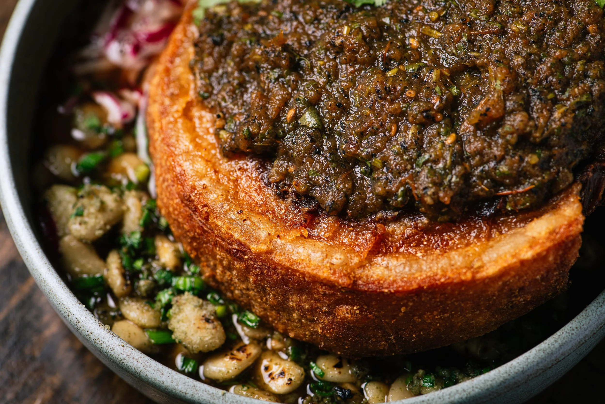 Close-up of a cooked pork shoulder topped with a thick sauce, sitting on a bed of cooked beans and chopped green herbs.