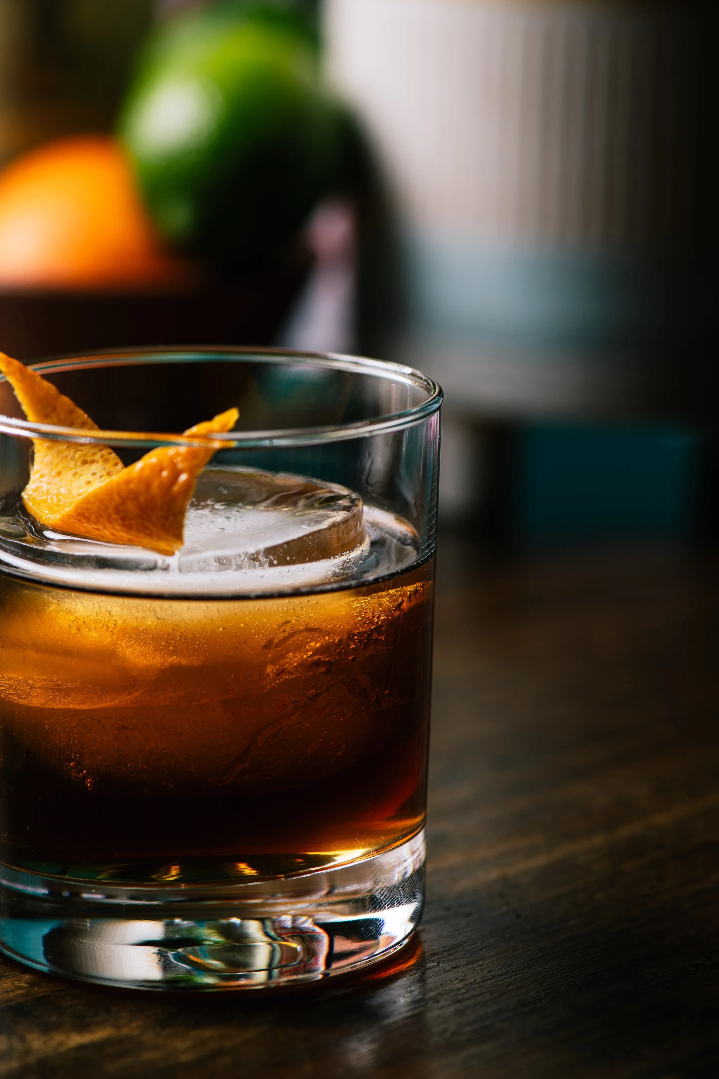 Close-up of a glass of dark cocktail with ice and a twisted citrus peel garnish on top, on a wooden surface with a blurred background including a green fruit and a bowl.