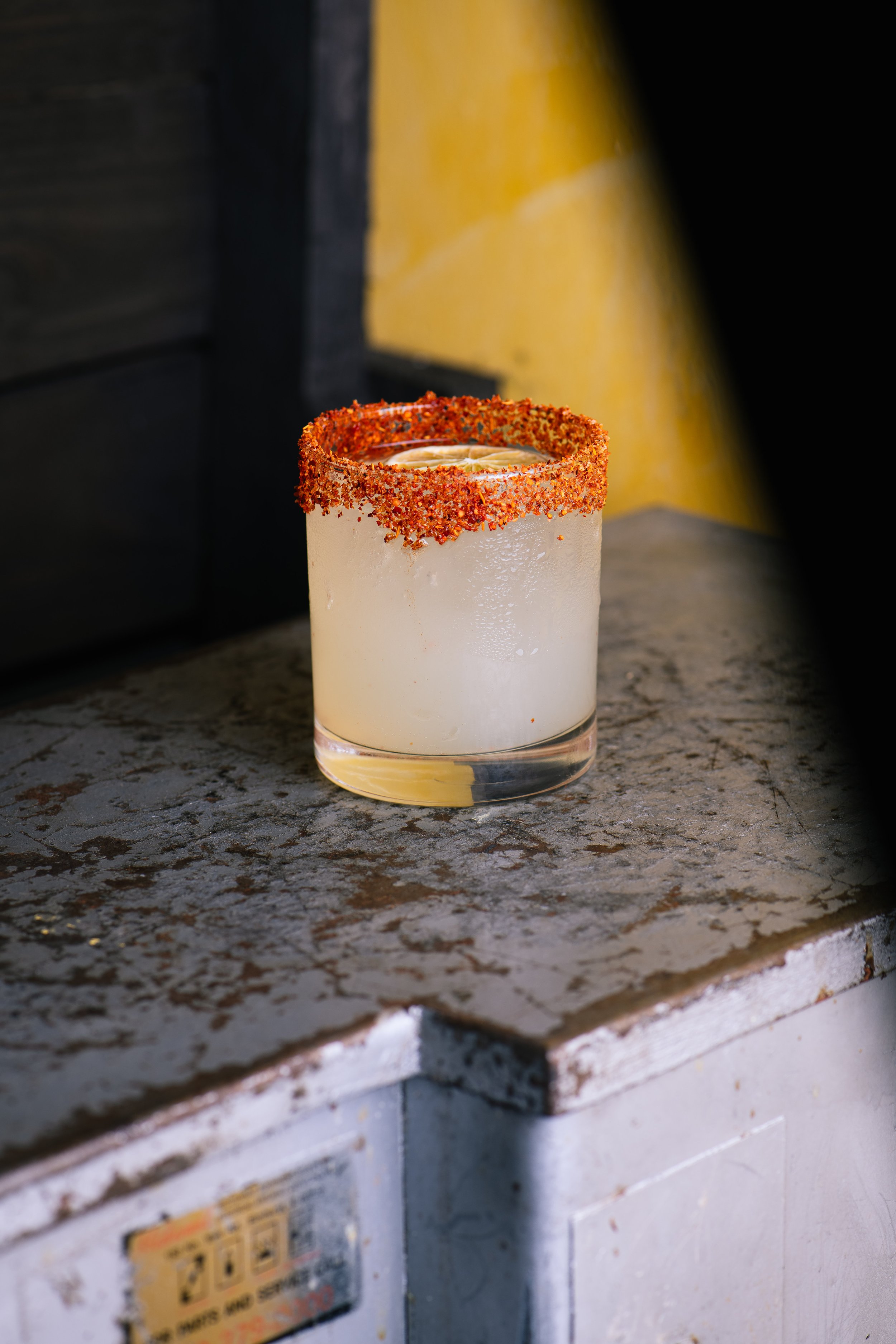 A glass cocktail with a chili rim on a rustic countertop, with a yellow wall and black furniture in the background.