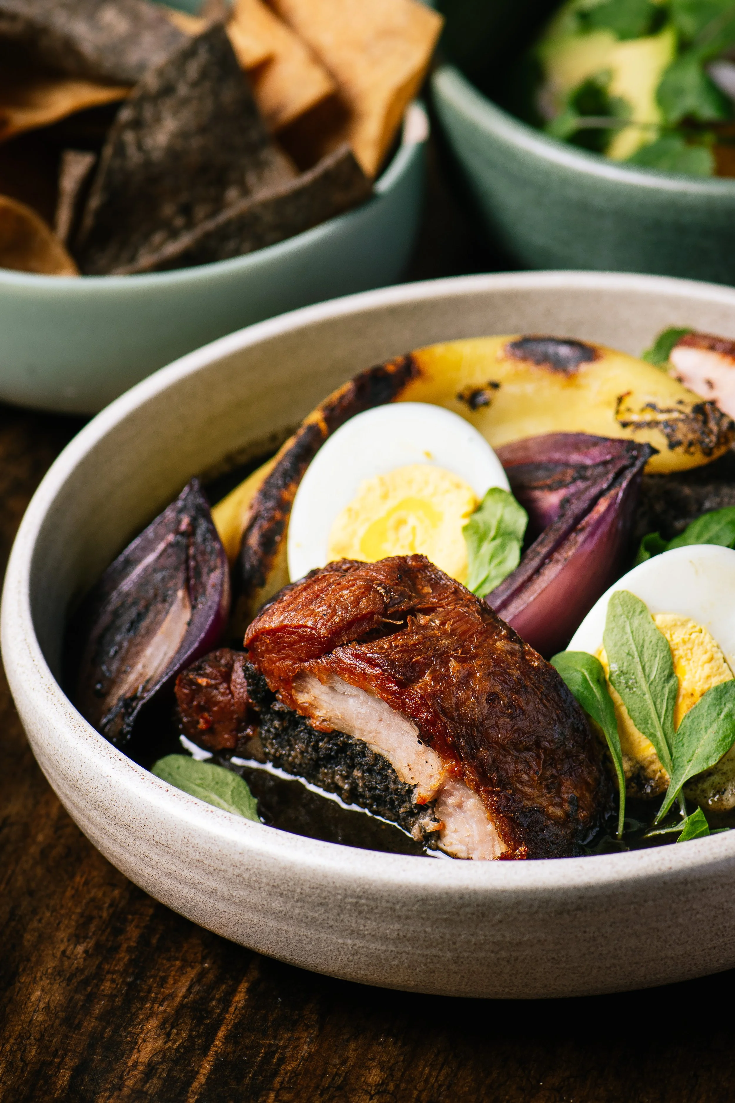 A bowl of food with grilled chicken, hard-boiled eggs, grilled eggplant, and greens.