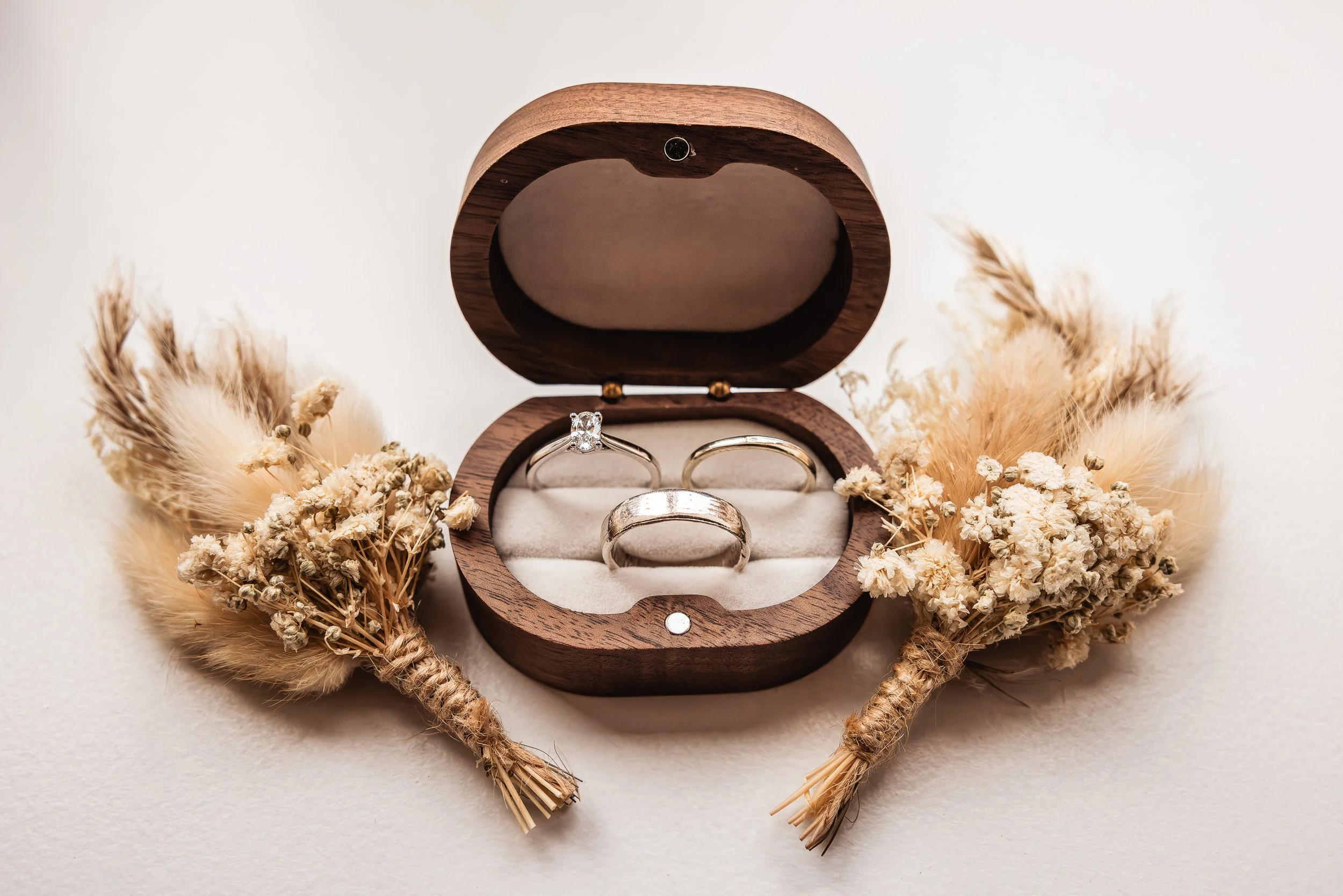 A wooden jewelry box containing three rings, surrounded by dried flowers on a white surface.