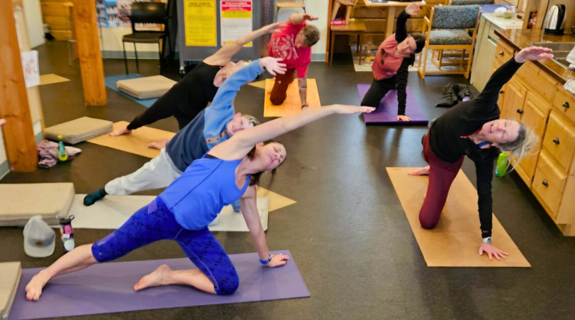 Group of adults practicing yoga in a room with wooden furniture, some on mats in various poses, stretching with arms extended overhead.