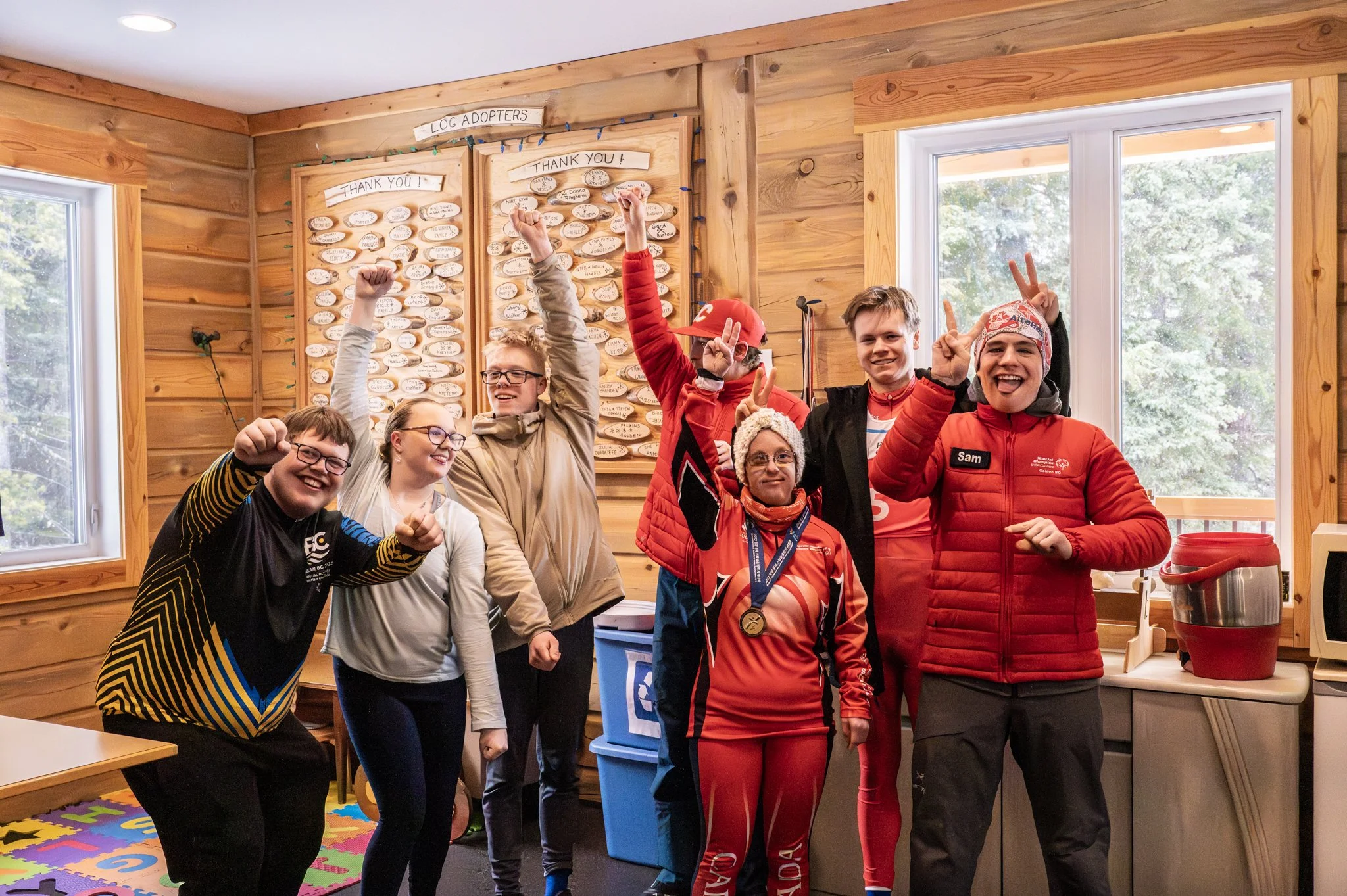 Group of people celebrating indoors with raised fists and smiles, in a room with wooden walls and colorful floor mat, relative to a decorated board that says 'Log Adopters' and 'Thank You'.