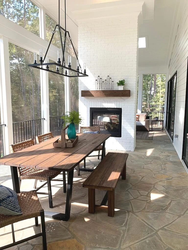 Screened porch with stone floor, dining table and chairs, double sided fireplace, chandelier and edison bulbs.