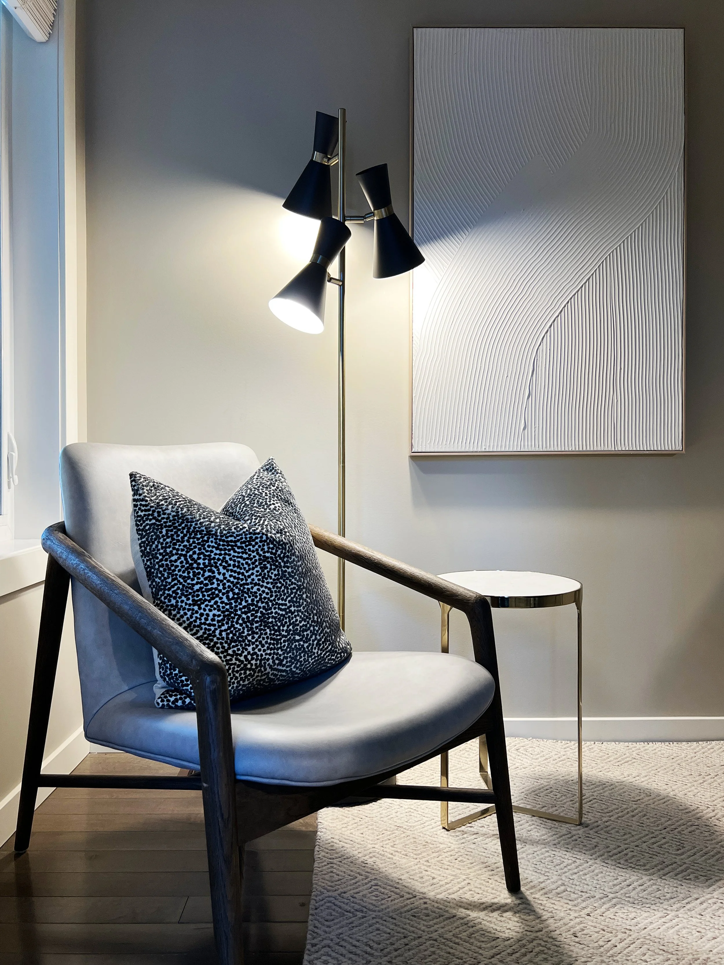 Living room corner with a white armchair, a black and white patterned pillow, a side table, wall art with textured white lines, and a modern black and gold floor lamp.