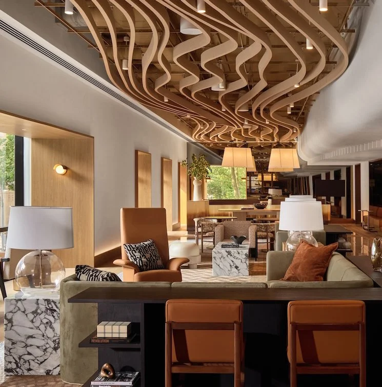 Interior of a modern hotel lobby with wooden wave-like ceiling design, stylish furniture, lamps, and large windows letting in natural light.