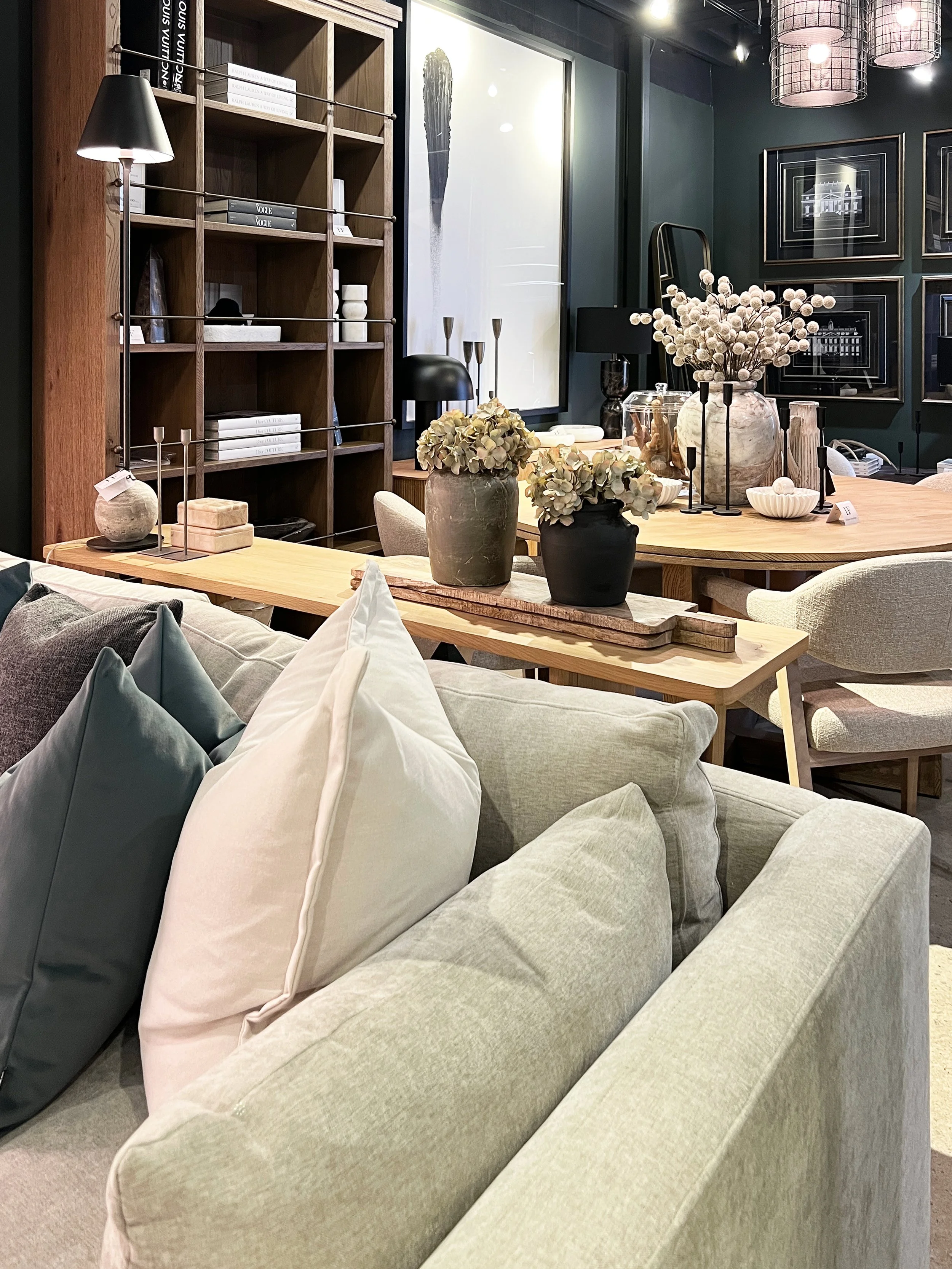 A cozy living room with a beige sofa in the foreground, adorned with various pillows in pastel shades. A wooden coffee table features vases with dried flowers and candles. In the background, a dark wall displays framed artwork, and there is a round wooden dining table with decorative vases and flowers. A bookshelf, lamps, and ceiling lights complete the decor.