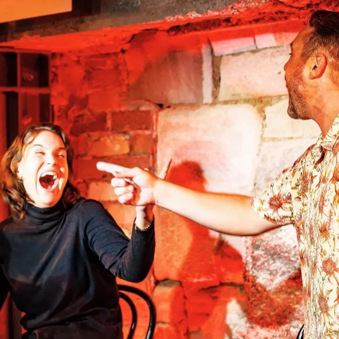Two performers point at each other while laughing delightfully.