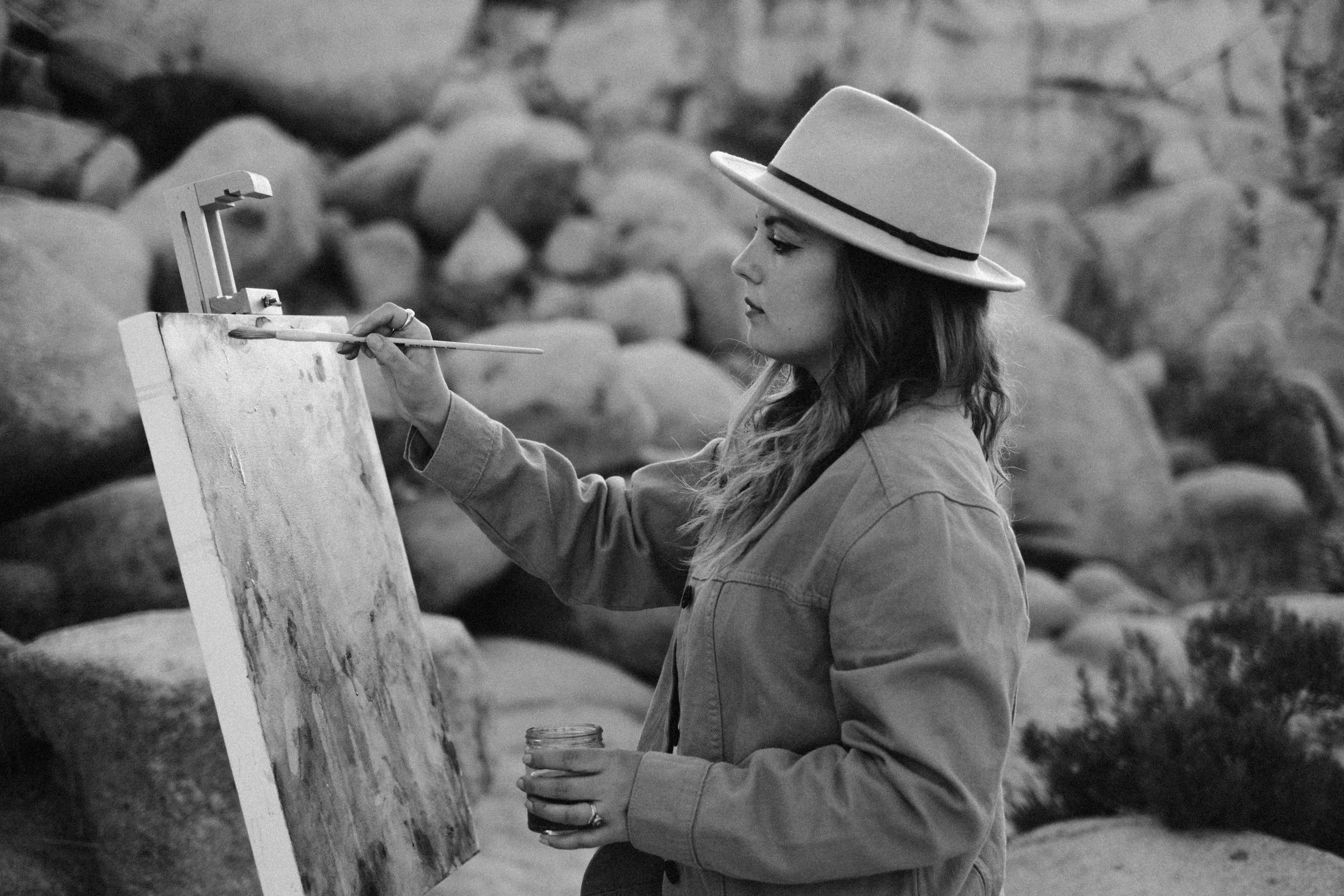 Woman painting outdoors with rocks in background, wearing a hat and holding a canvas.