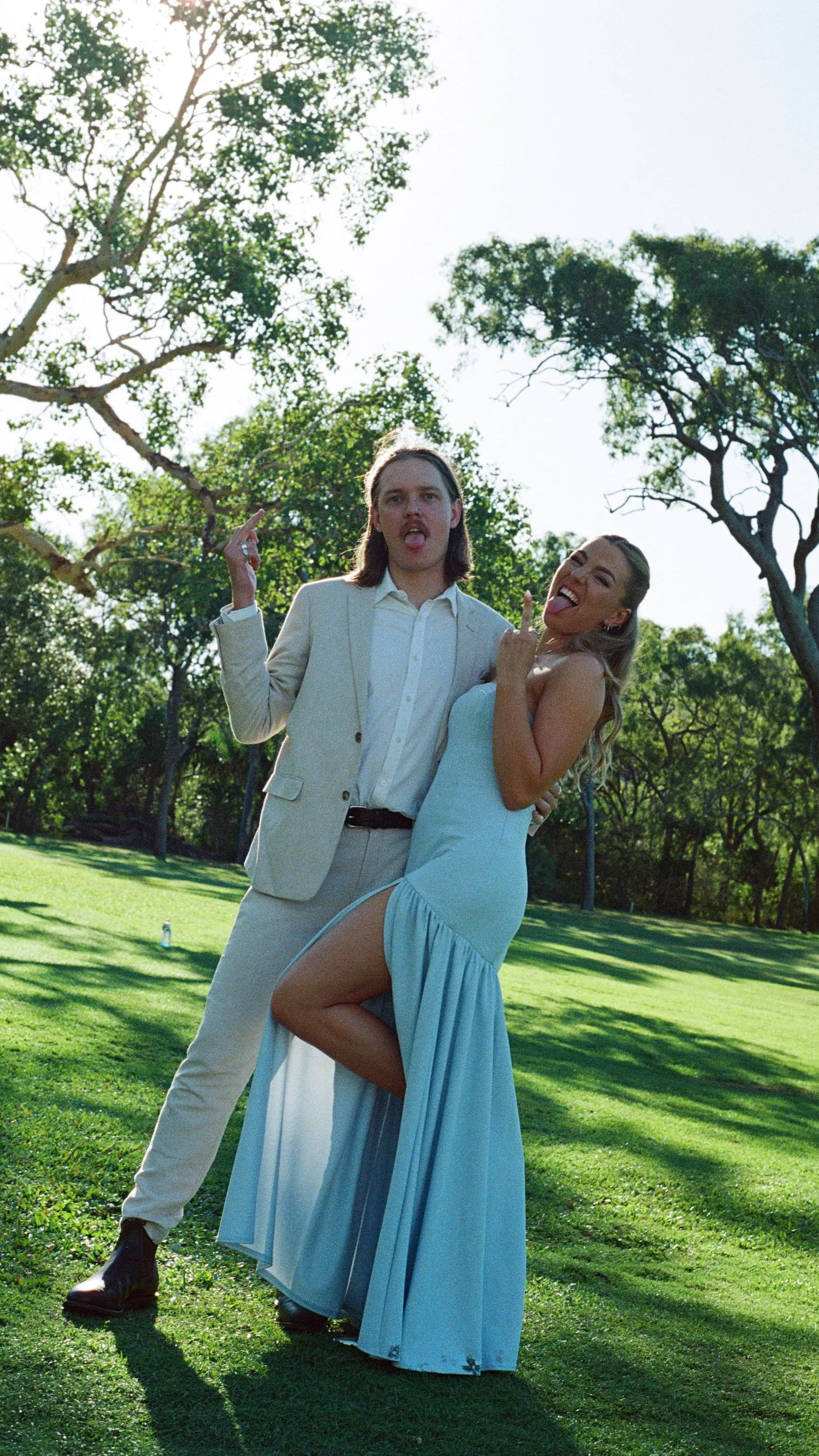 A couple stands outdoors on a sunny day, both making playful facial expressions and gestures, with trees and grass in the background.