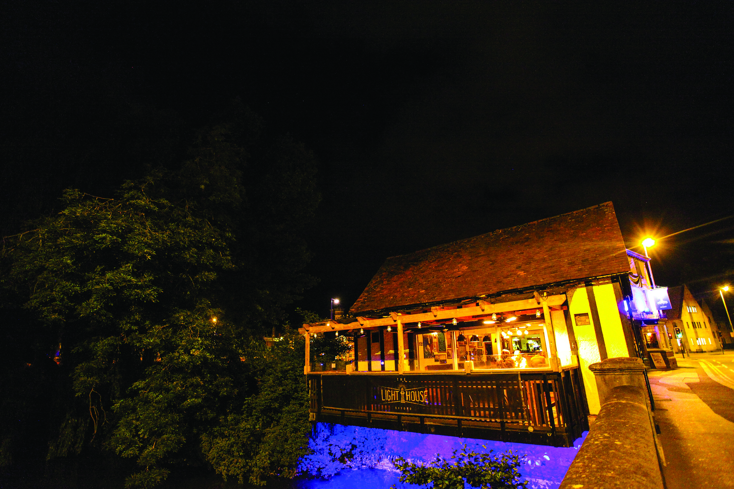 A image at night time of the balcony at the lighthouse which is overhanging a river and has a blue light shining down underneath.