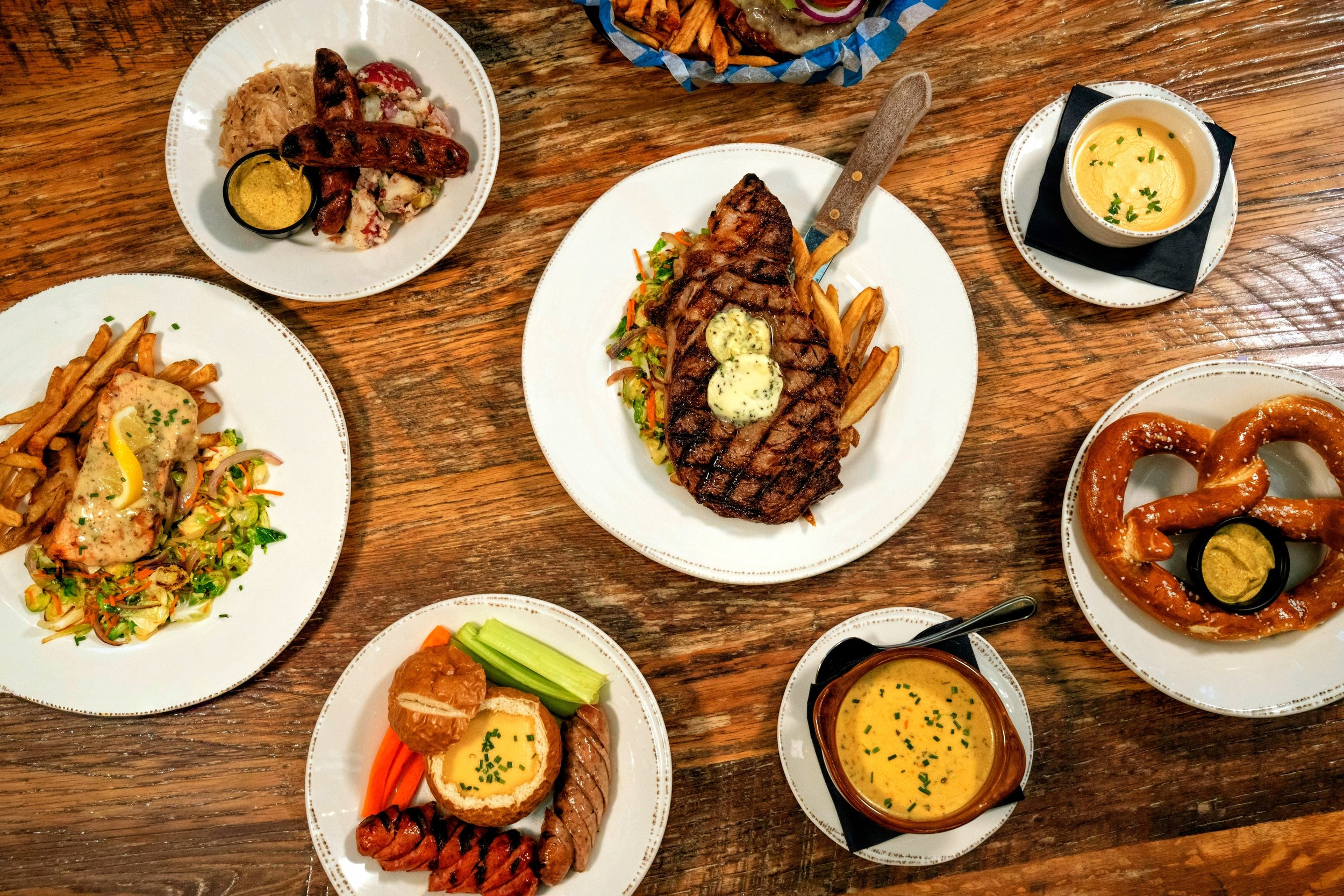 A group food photo of ribeye steak, salmon, wursts, pretzel, and bier cheese.