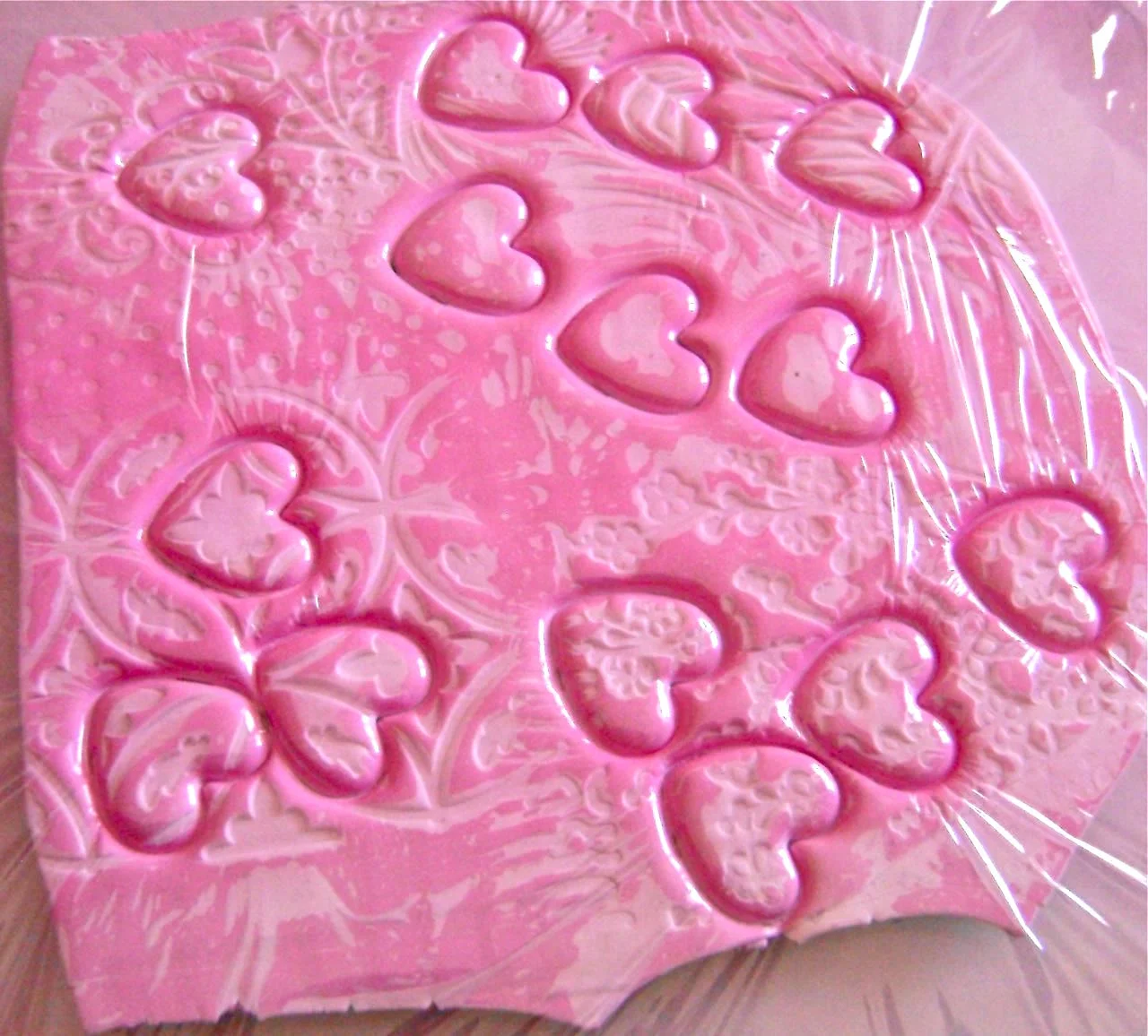 step 4 - Place kitchen plastic wrap over the clay sheet (mine is pink) and press the heart clay cutter down over the designs to capture details that appeal to you. The plastic wrap will create nice rounded edges. There is no worry of mistakes here. If you don’t like what you’ve done just roll the clay together and start over.