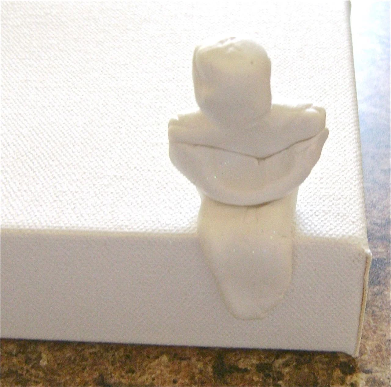 step 2 - Roll another piece of clay to drape across the front. Press it to the sides to create elbows and folded arms. Bend the mummy into a seated position. Press it to the edge of a 90 degree angle to be sure it is balanced and will sit without falling over.