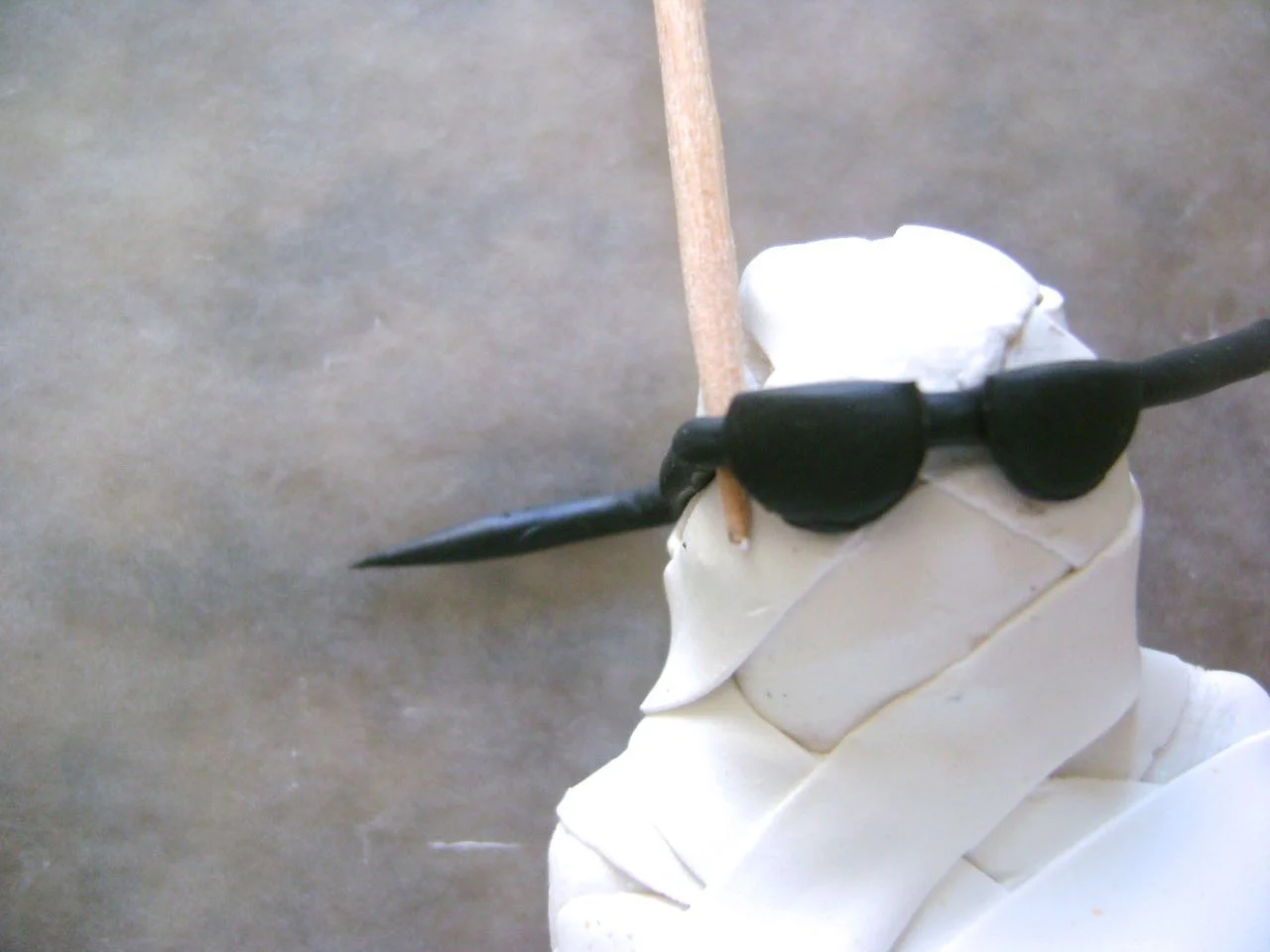 step 8 - Position the glasses on the mummy. Place a toothpick against the corner of the glasses and wrap the arm back to the ear area. Cut away excess black clay and press the arm to stick it to the head.