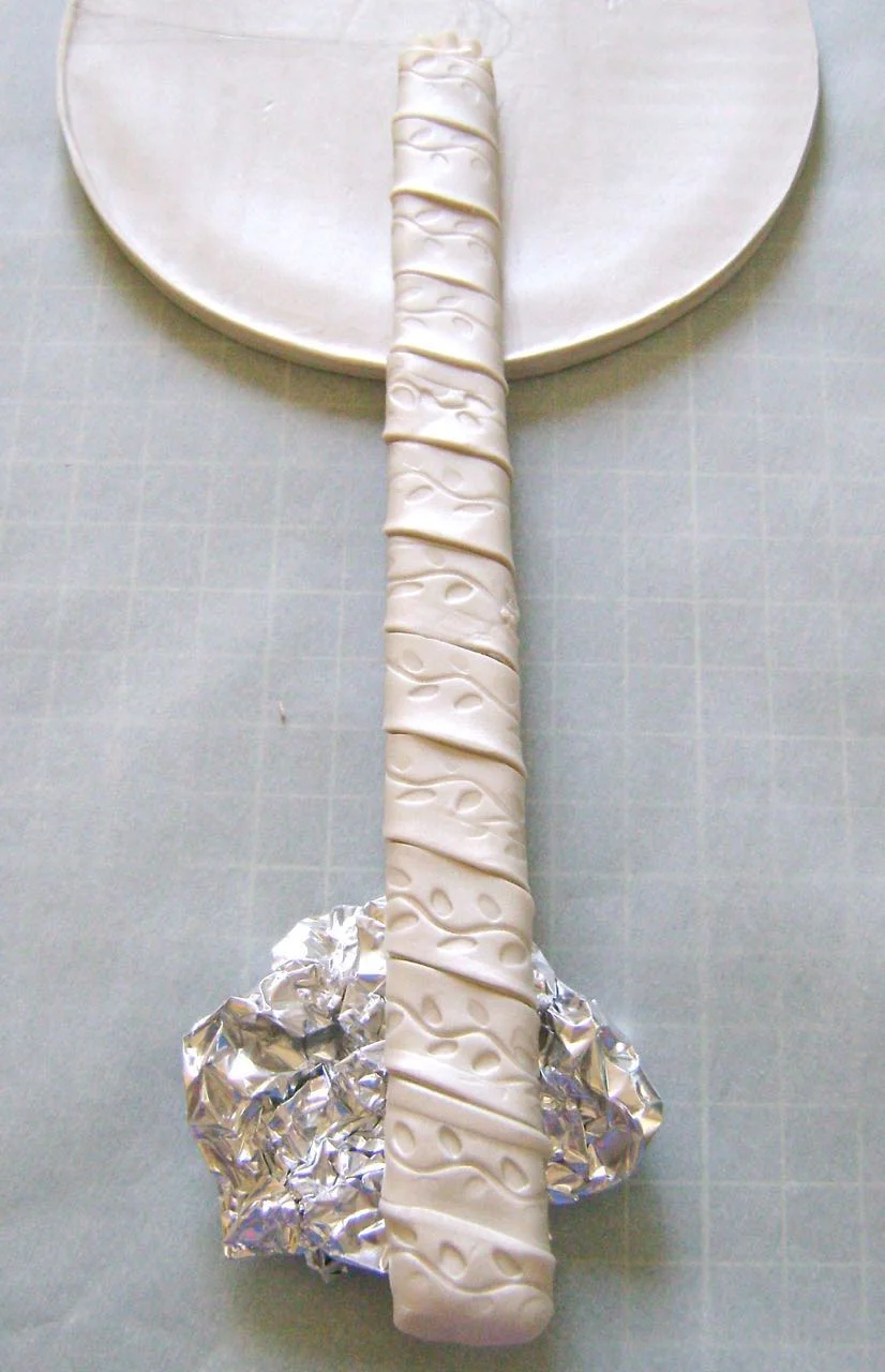 step 10 - Flip the clay base onto a baking sheet lined with parchment paper, textured frame face down. Press the baked handle against the center of the round shape. Place crumpled aluminum foil under the handle to keep it horizontally level with the base.