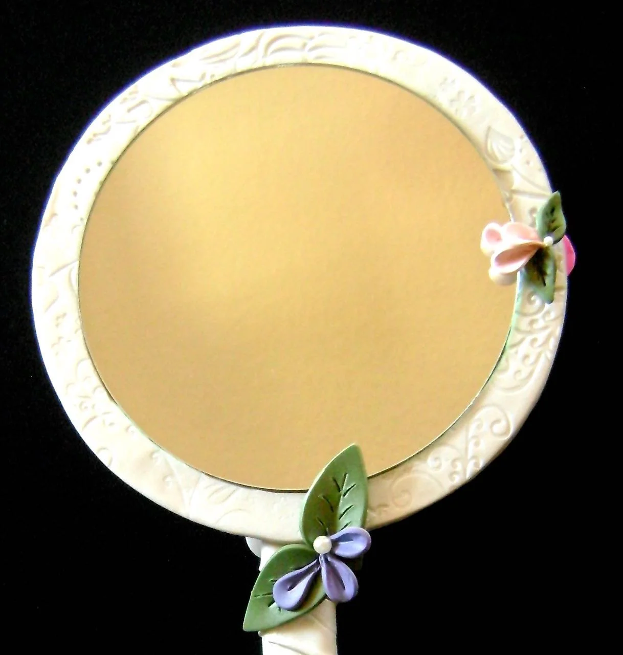 step 17 - Remove the cushion pads on the backside of the mirror. Use glue to attach the mirror inside theclay frame. Create a few more petals and leaves to attach to the frame. Bake them as the others. Once cool, attach pearls and glue the flowers to the frame slightly overlapping the mirror’s edge.