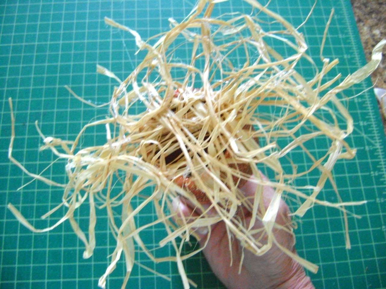 step 14 - Lay out a clump of raffia on a protected work surface. Apply hot glue to the bottom perimeter of the pumpkin. Press it into the raffia. Repeat 2 or three times. Trim the raffia as desired. Turn the pumpkin over and cut out the raffia covering the center hole. Cover an LED votive light with the pumpkin and turn on the switch.