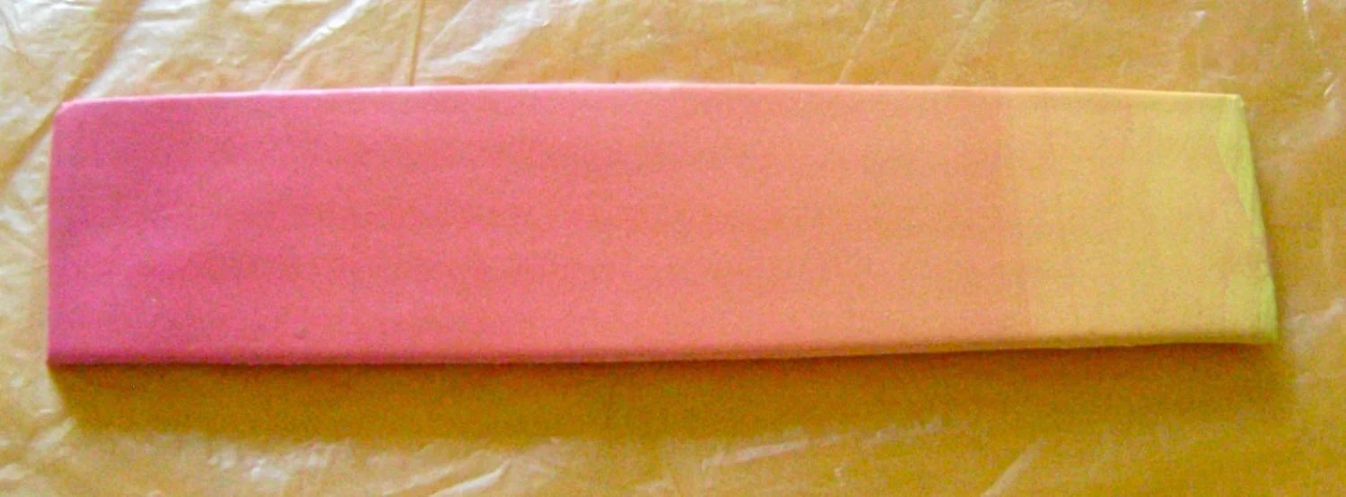 step 4 - Fold the blended clay in half along the length (yellow to yellow edge and pink to pink edge). Feed it into the pasta machine on #4. This will lengthen the piece and soften the look of the blend. Cut the strip to measure 1” wide.