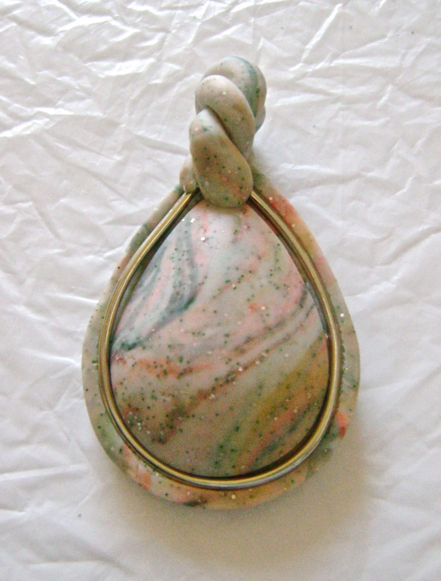 step 12 - Roll a small piece of the excess clay in your hands. Fold it in half then twist the clay together. Slice one end at a 45 degree angle so the clay will be angled under when in place. Press that end onto the edge of the cabochon and bezel. Curve the twisted clay into a loop to create a bail for the chain and slice the other end of the clay at a 45 degree angle. Leave enough of a space in the loop of the bail for the chain you plan to use. Press the free end of the clay to the back of the bezel. 