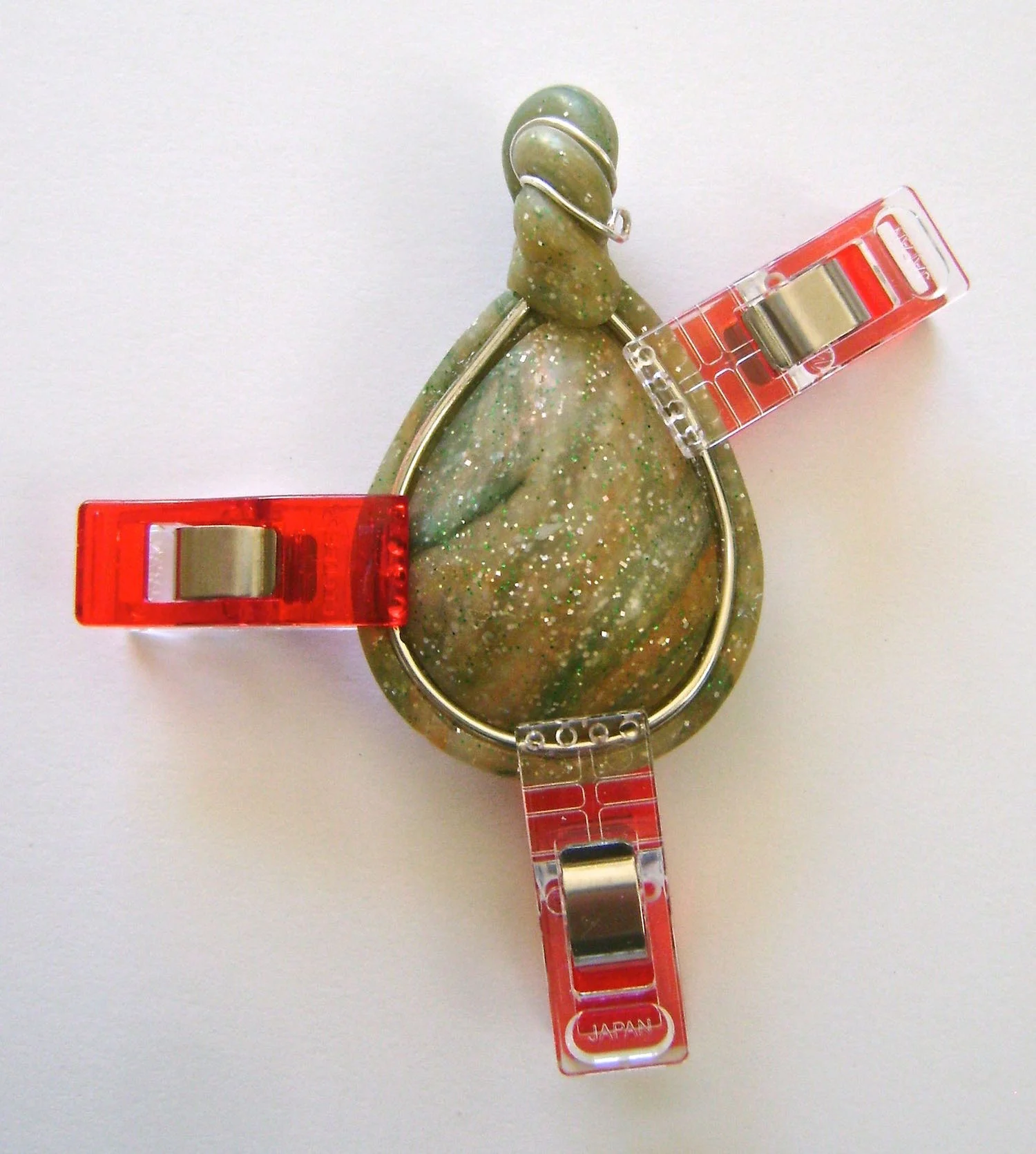 step 18 - Using a toothpick, apply adhesive to the gutter between the cabochon and the bezel. Carefully insert the ends of the teardrop shaped wire under the bail. Press the rest of the wire into the gutter. Hold it in place with clips and allow the adhesive to set overnight.