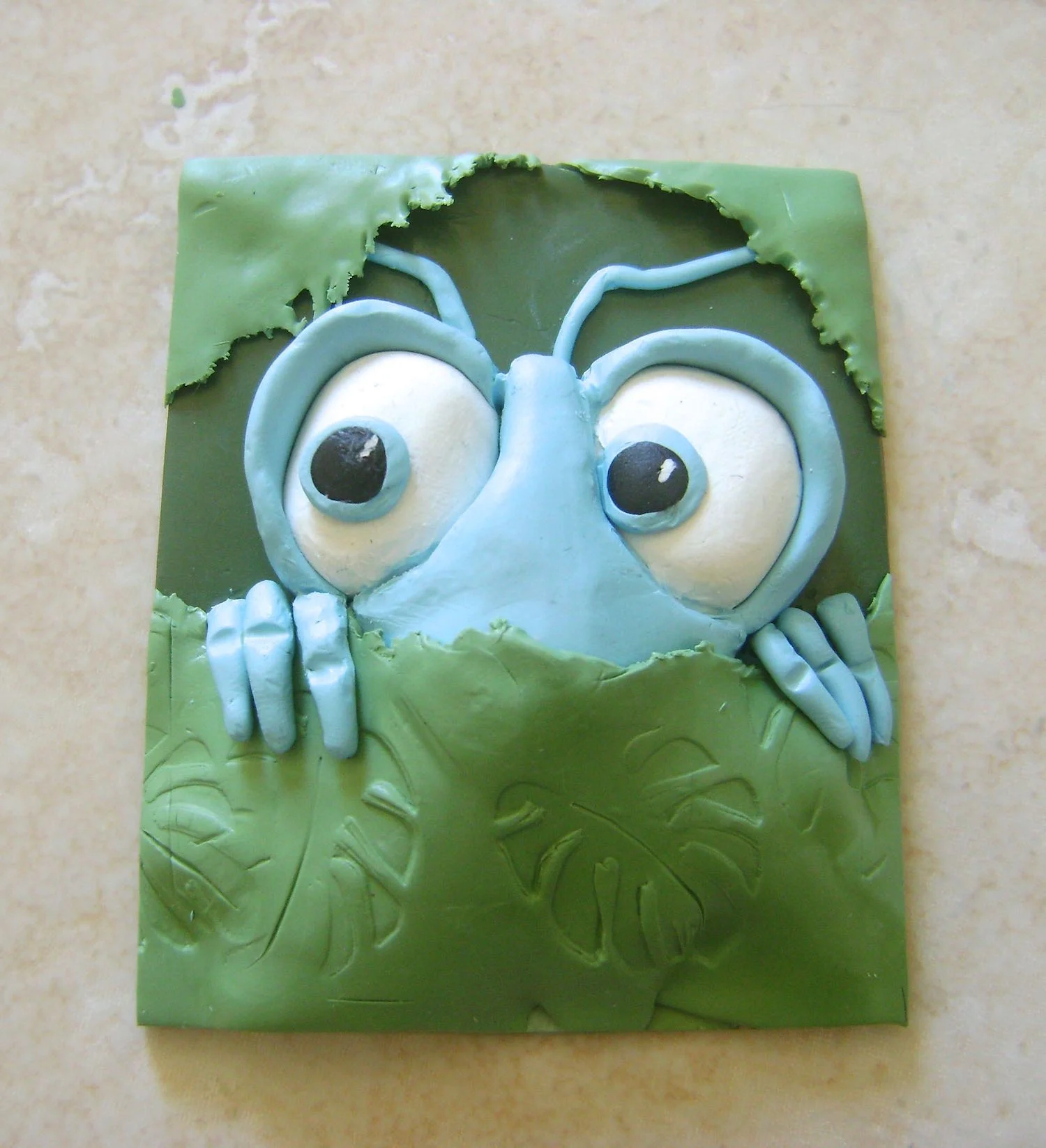 step 7 - Tuck one end of the fingers behind the ragged leaf. Press the fingers in place. On the work surface, roll out thin tubes for antenna and position them on the dark green base. Create two more ragged clay pieces for the leaves at the top of the project. Gently press all four edges of the frame to bond the layers together. Use a tissue blade to cut the excess clay to match the base size. Pinch the frames of the eyes and bend them away from the face slightly.