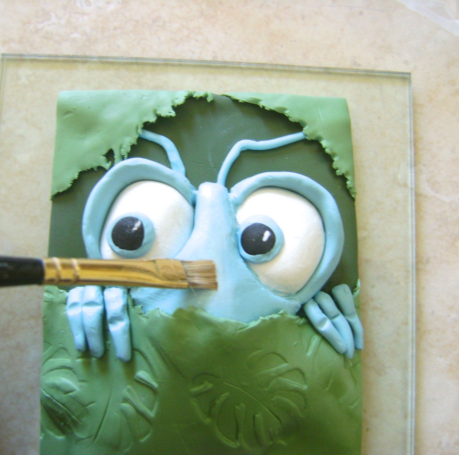 step 8 - Smooth sharp edges and nicks in the clay with your finger where necessary. Add details to the center blue clay with white pearl Mica Powder applied with a brush. With a thin brush, paint the inside of the eye frames, the lower eyeballs, and the depressions of the fingers with a mix of blue and black acrylic paint. Make a wash of copper paint and brush it on the leaves. Wipe away the surface color leaving the paint in all the crevices to accentuate the leaf shapes.