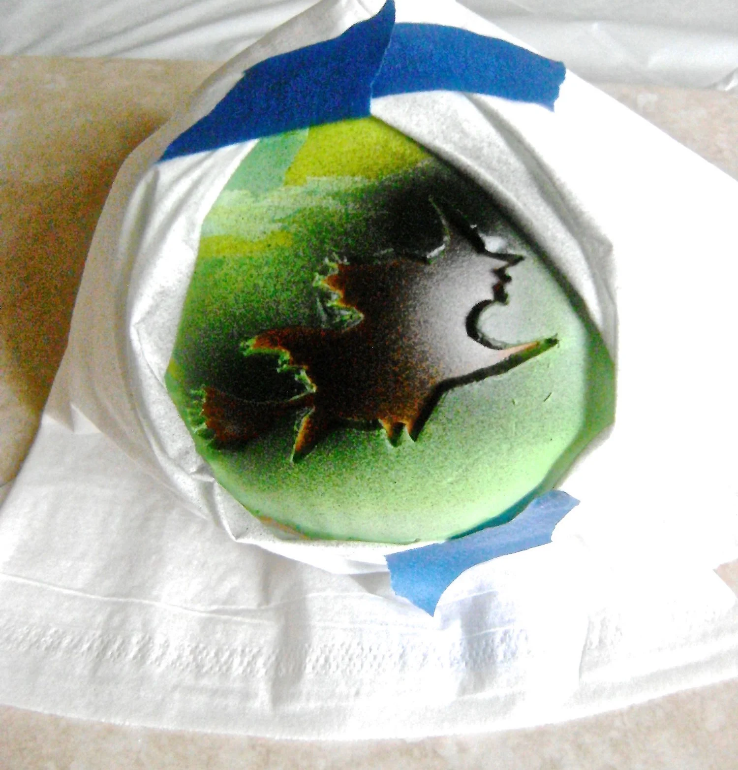 step 13 - Press the ornament onto another piece of scrap clay on the work surface. This will hold the ornament in place while you work. Warp the clay stencil around the ornament. Mask off surrounding areas. Airbrush the stencil with black paint. Remove the stencil, reposition it on the ornament two more times to create a total of three witches.