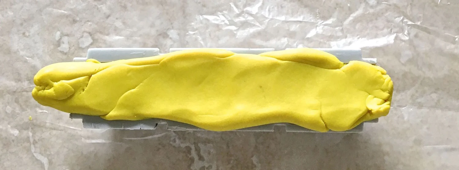 step 1 - Roll out yellow clay on the work surface to create a tube. Mist the inside of a 18mm Sculpey Bead Maker. Press the tube into the opening.