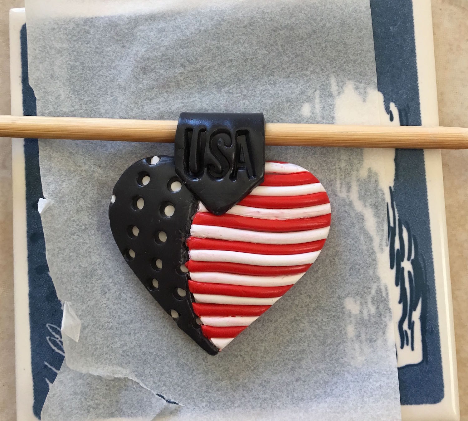 step 15 - Press the point of the clay to the center of the heart. Bring the rest of the strip to the back of the pendant and trim away the excess. Place a wood pick into the bail loop to hold the strip open while baking.