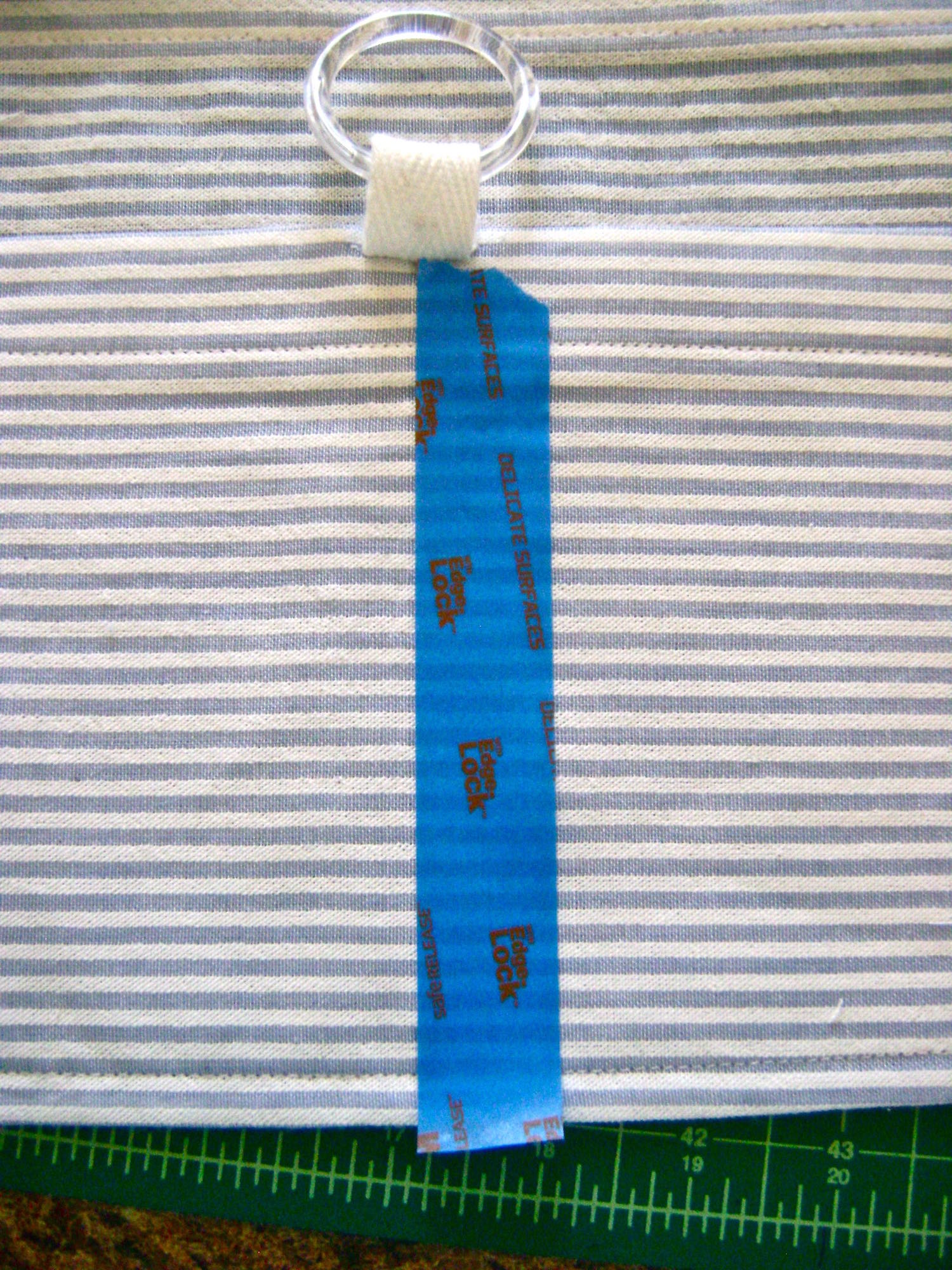 step 18 - Slip a piece of cotton tape through the plastic ring. Sew the tape to the top center of the pocket with the raw edges inside. Mark the vertical center of the pocket with tape and stitch along the tape edge from the bottomup to the tape to create a divided pocket.