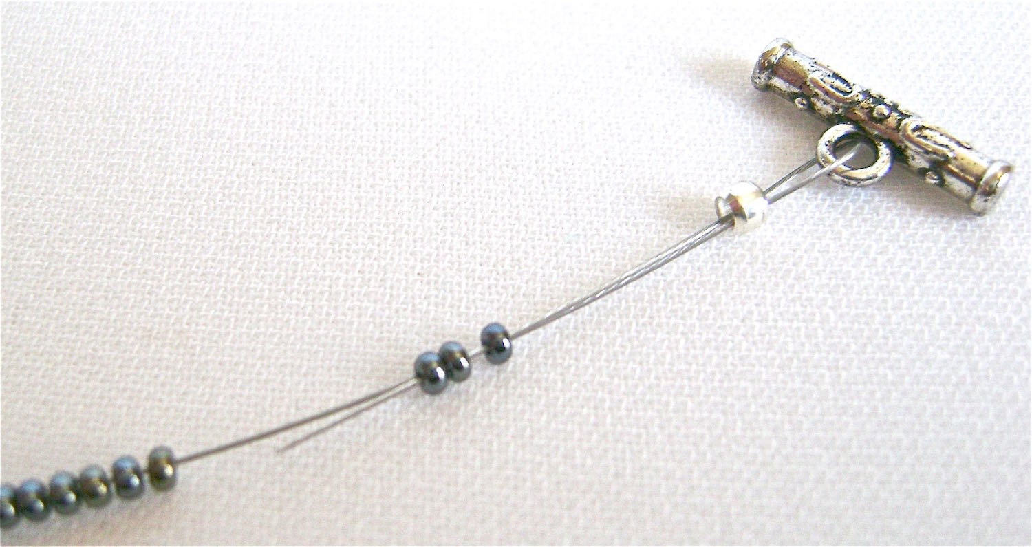 step 11 - Add one end of the clasp onto the wires then bring the wire ends ends back through the crimp bead and if possible, through a few seed beads. Pulling on both wires simultaneously, slide the crimp close to the clasp and secure with a double crimp. Trim the excess wire and slide all the seed beads over it. Remove the tape from the other two wire ends and attach the clasp as described in step 10 and 11.