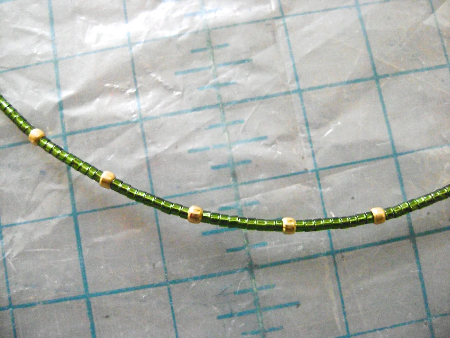 step 14 - Make the necklace by cutting a piece of gold wire to 16”. Add beads as follows: 2- oval gold beads, 1- round gold bead, 18 - green seed beads. Add nine sets of 9 nine green beads and by one round gold bead. Finish with 18 green beads, one round gold bead and two oval gold beads to match the other side.