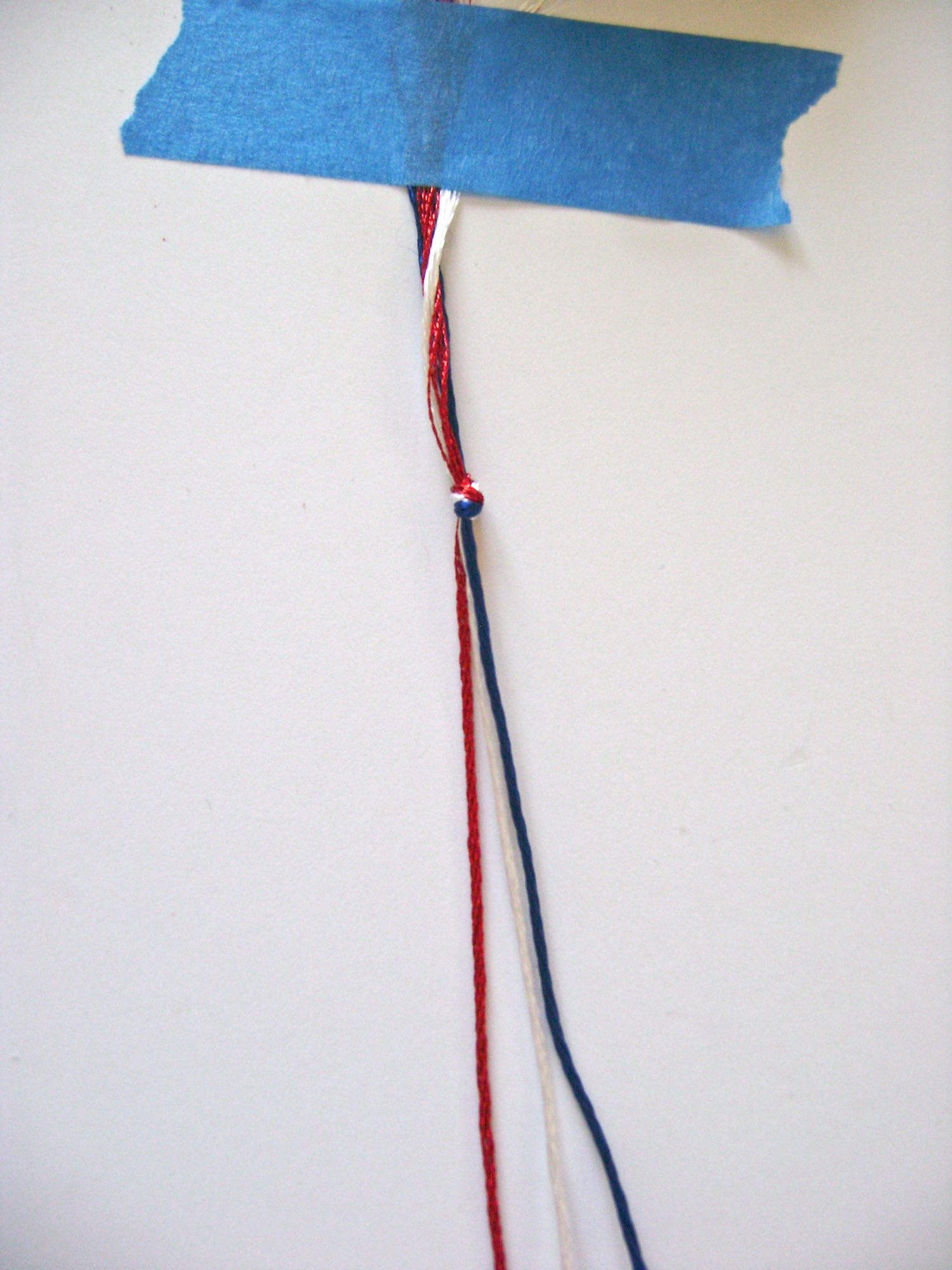 step 5 - Gather three strands of embroidery floss, red, white, and blue and knot them together. Tape the knotted end to the work surface.