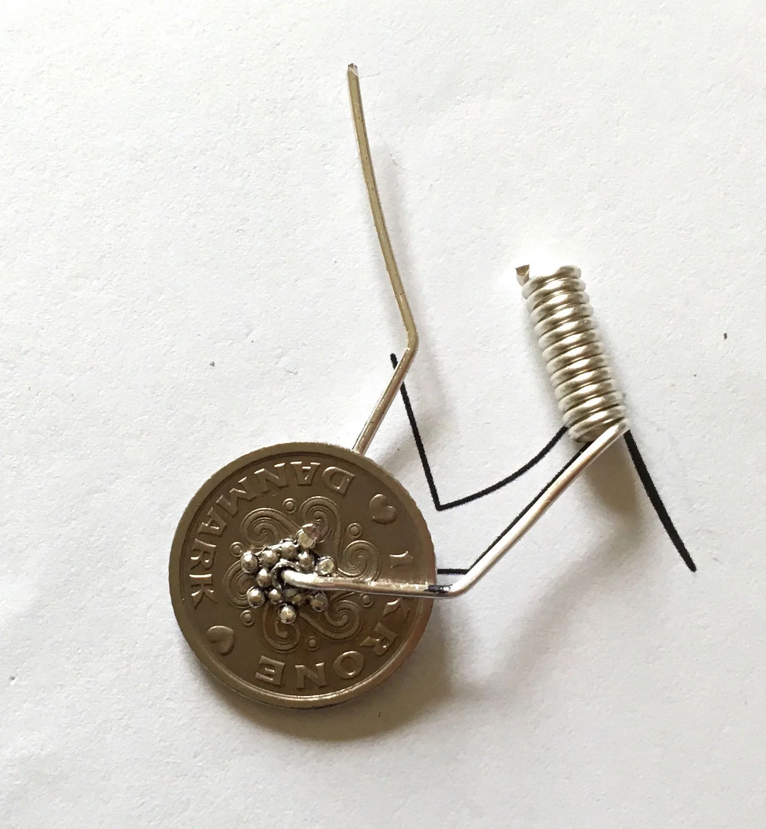 step 5 - Center the coin over the second bend mark. Squeeze the beads flat against the coin and press the wire flat and upwards on both sides. Use flat nose pliers to bend the wire at an angle at the top end of the template line.
