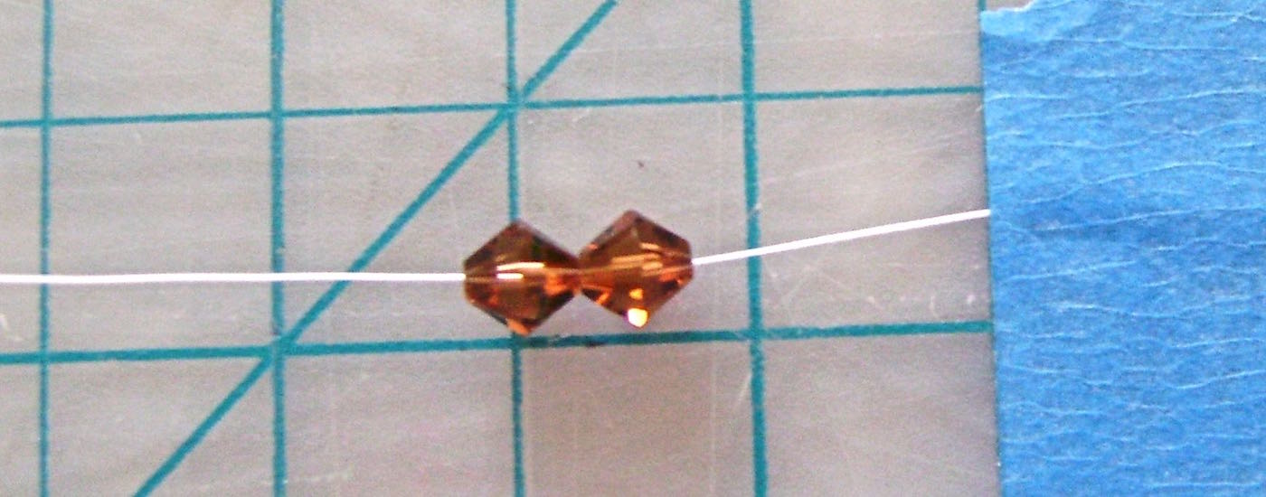 step 1 - Thread a big eye needle with 3 yards of thread. Put the needle through two 6MM beads. Pull through leaving a 10” tail. With painter’s tape, secure the tail to the work surface. Two beads are added to the thread for the first step only. Steps that follow only require the addition of one 6MM bead at a time.