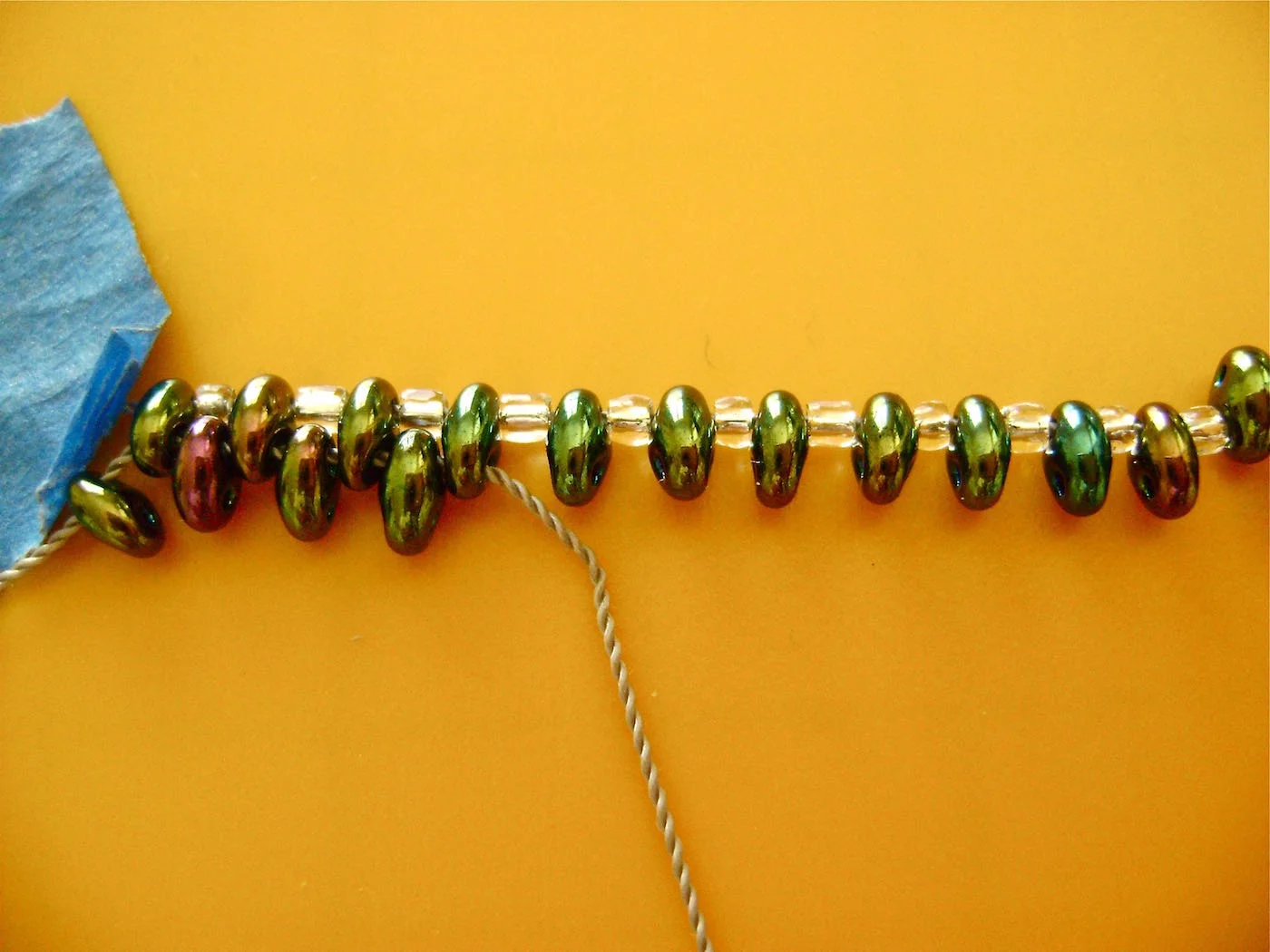 step 3 - Start the second row with a new strand in the beading needle. Tape the end to the work surface. Put the needle through the bottom hole of the first bead in the first row. Add a new bead. Put the needle through the bottom hole of the next bead in the first row. Continue working across the row in this way. At the end of the row, remove the needle, tape down the thread and add a new strand to the needle. (You may note in the photos that I started the bracelet with cord. I stopped when I realized it would be too thick for this project and started over again using thread.)