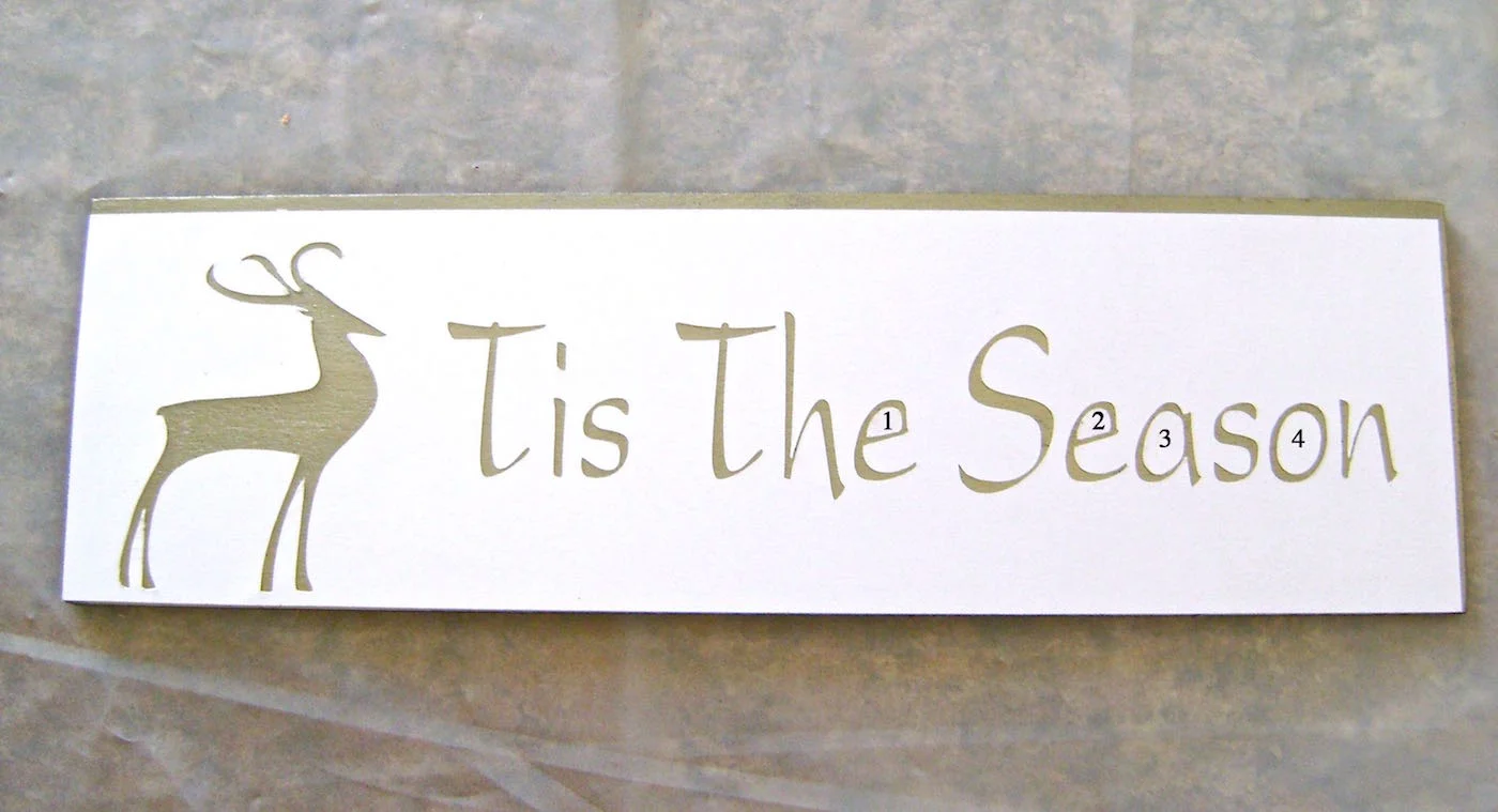 step 6 - Spray the back of the stencil and the cut pieces with adhesive. Position the stencil on the wood piece. Place the cutouts inside the corresponding letters sticky side down. Check to be sure all fine edges adhere to the wood. Airbrush the stencil with brown paint.