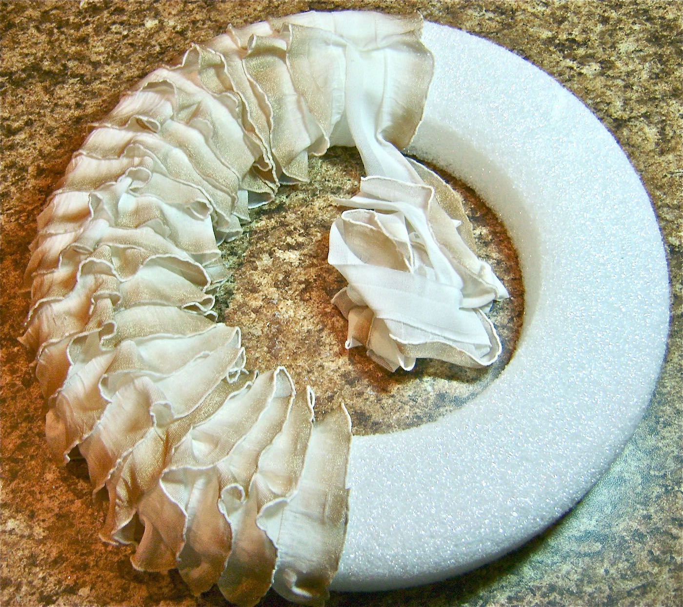 step 7 - Continue working down the wreath until you reach the bottom center. As you work the curved areas you will need to overlap the ruffles at the inner edges of the wreath more than the outer edges because of the difference in diameter. When you’ve finished adding ruffles to one side of the wreath, move to the top center and attach the end of a ruffle strip to the backside of the wreath. Overlap the strips in the center which will create a flat double border to be covered later. Repeat steps 5-7 above to finish covering the wreath with ruffles.