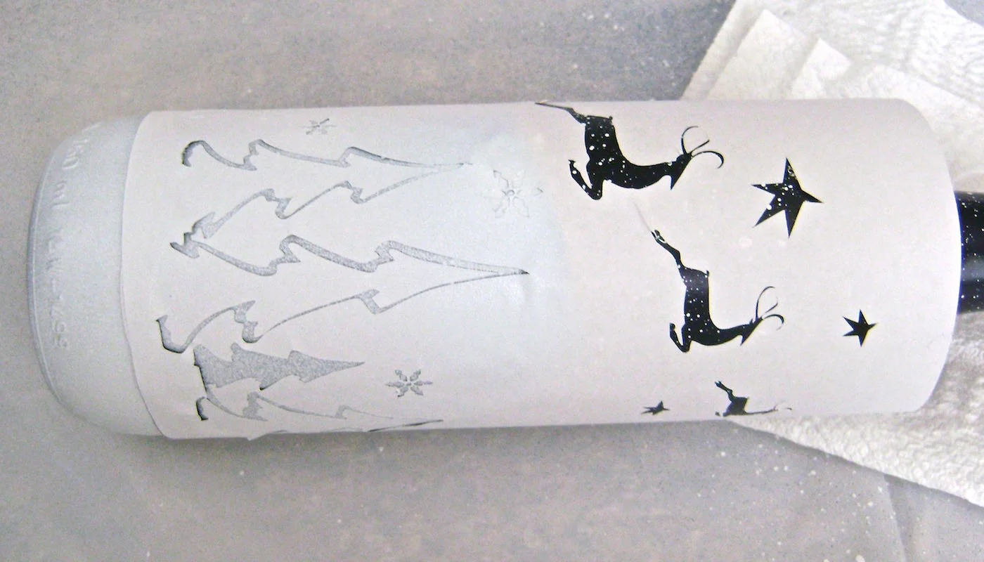 step 6 - When the paint is dry, remove the backing from the stencil sheet for the trees and snowflakes. Press the stencil onto the bottle. Align the bottom of the trees with the rubber band. Cover the reindeer and stars and airbrush the trees and snowflakes with white paint.