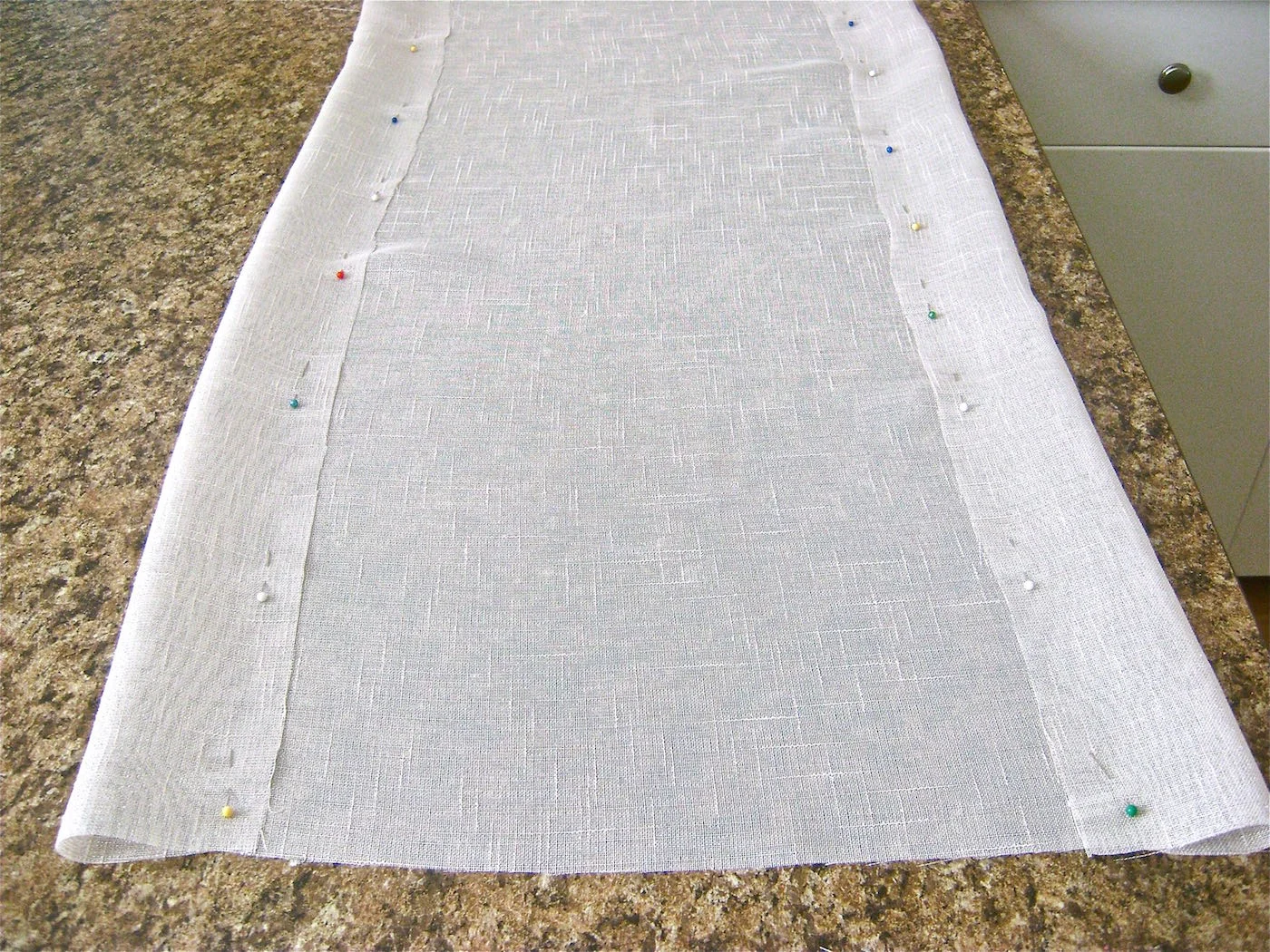 step 1 - Cut the length and width of the runner you need for your size table. The short finished ends should drop a minimum of 4” down from the table edge. I made my table runner 18” wide so that plates on both sides of it would not overlap the fabric or design. For an 18” runner, start with raw fabric cut to 24” wide. Fold and pin three-inch hems along each side.