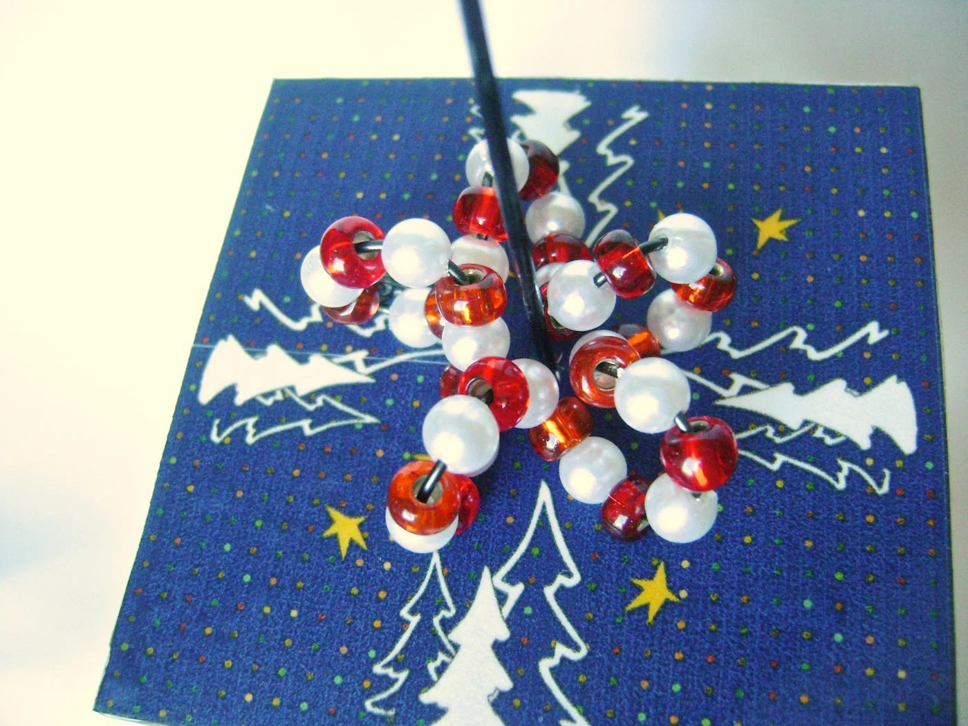 step 10 - The Beaded Trim With round nose pliers, create an open loop at the end of a ten inch black wire. Wrap the wire around a magic marker to create a spiral. Stretch the spiral enough to allow beads to move across the wire easily. Alternate red beads and white pearls. Squeeze the spiral together to make it the right size to wrap around the cube wire in a circle. Create a loop on the other end of the bead wire and attach it to the first loop. Squeeze the beaded wires together to create a star shape at the base of the cube.