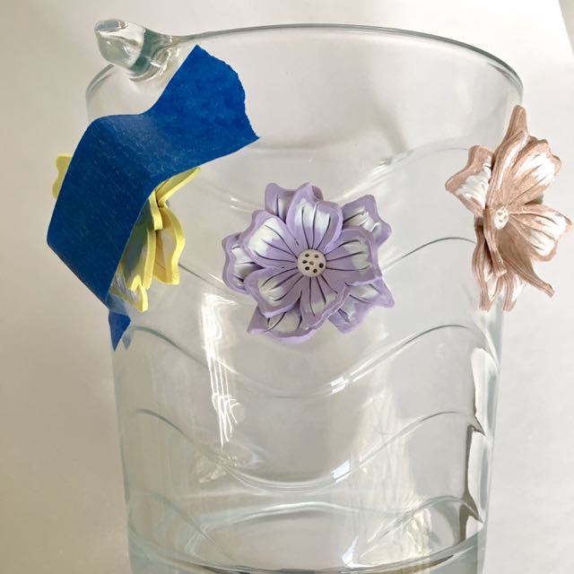 step 13 - Attach 8 flowers to the container with E6000 adhesive. Alternate the sizes in this top row as well as the colors. Apply masking tape to prevent the flowers from sliding down the glass. When the adhesive is set, remove the tape and apply a second row, placing these flowers slightly below and between the flowers in the top row.